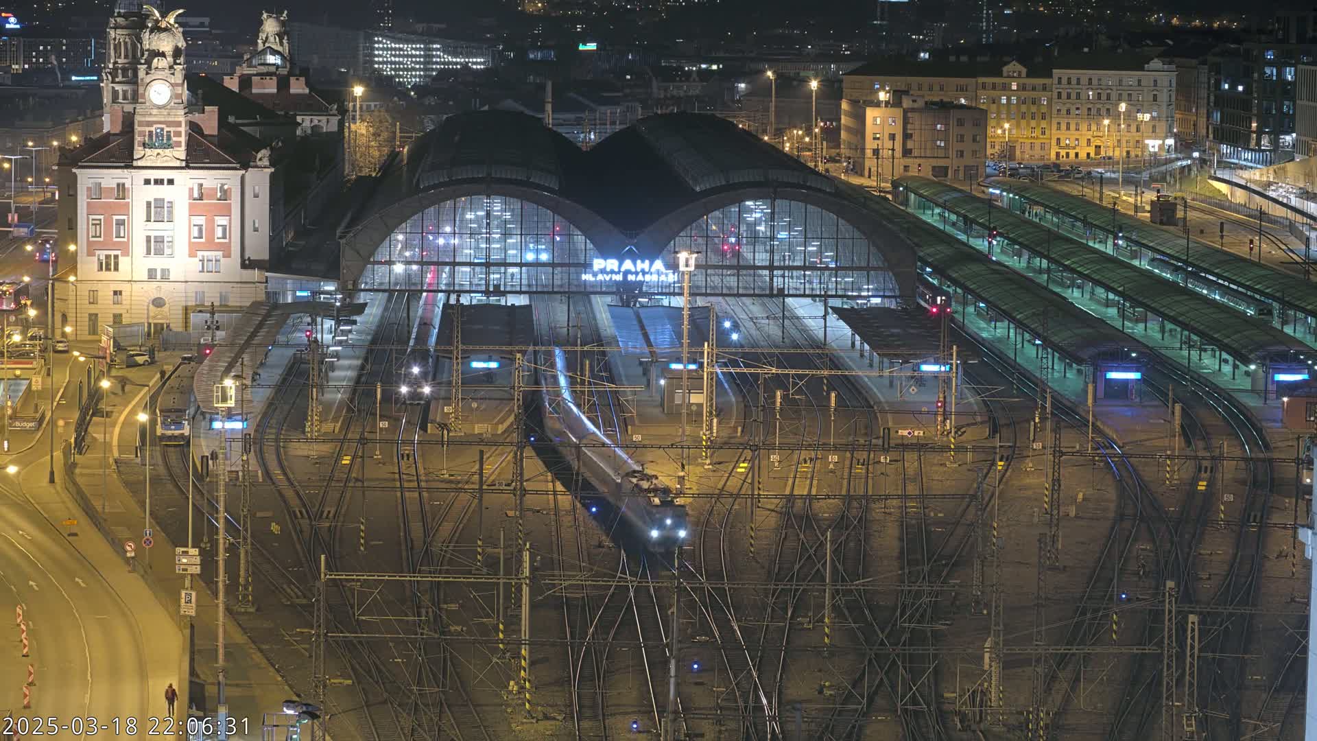 A high-angle nighttime view of a bustling train station with a train arriving at its platforms, surrounded by city buildings.