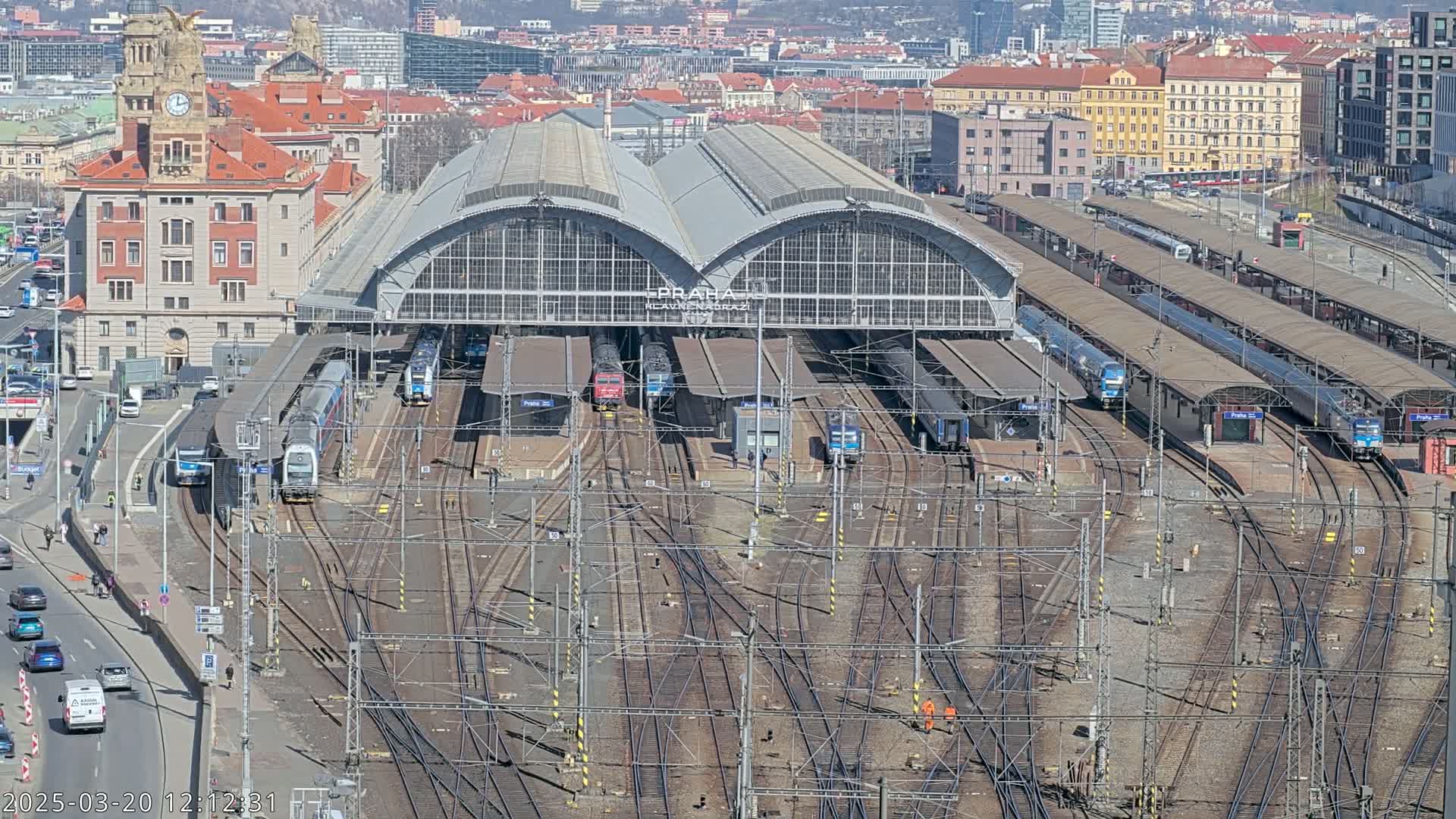 A large train station with numerous trains at platforms and a complex network of tracks, under a sunny sky.