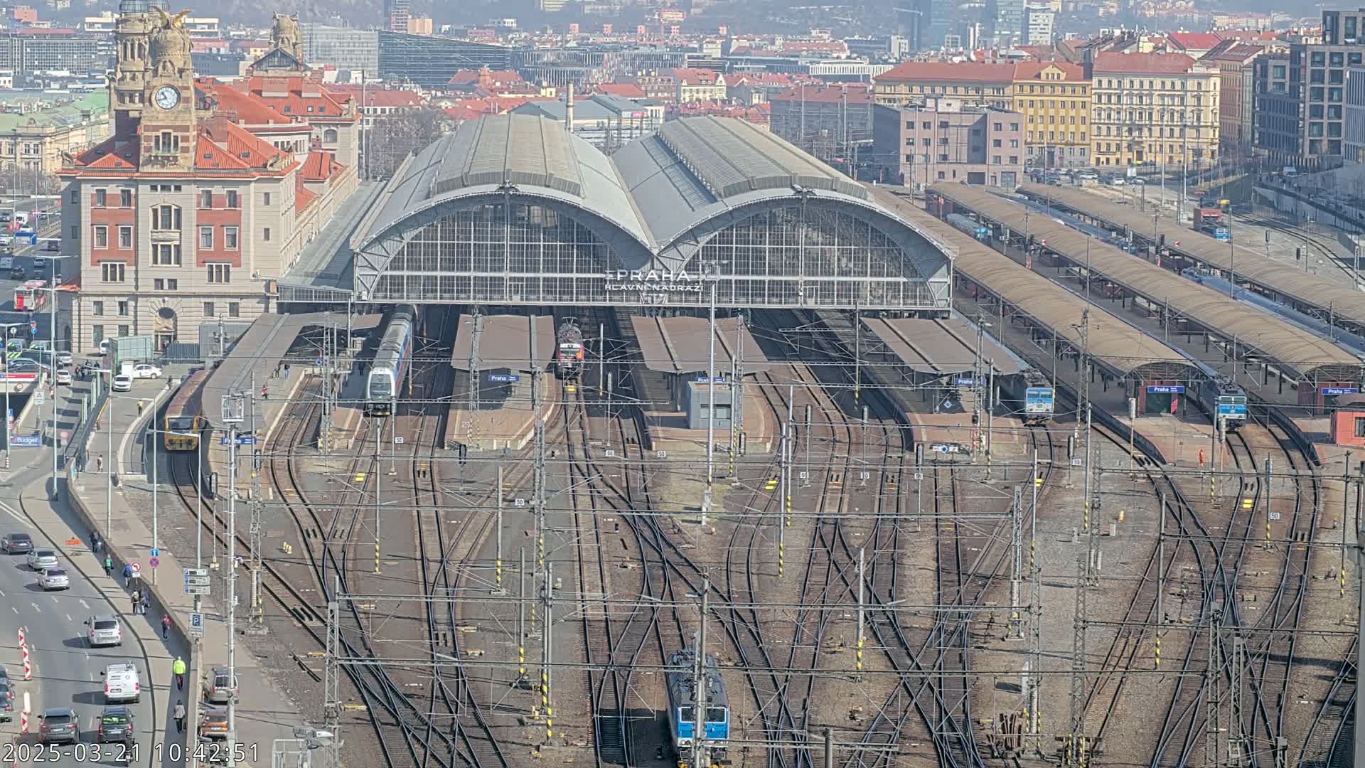 A large train station with many tracks and trains, under a sunny sky, is viewed from above.