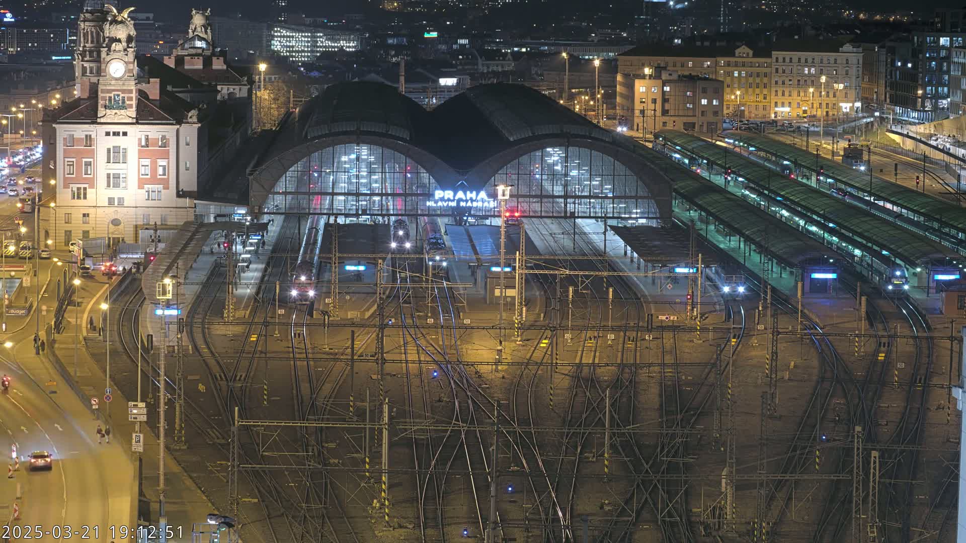 A nighttime aerial view of a large, illuminated train station with many tracks and platforms, surrounded by city buildings under clear skies.