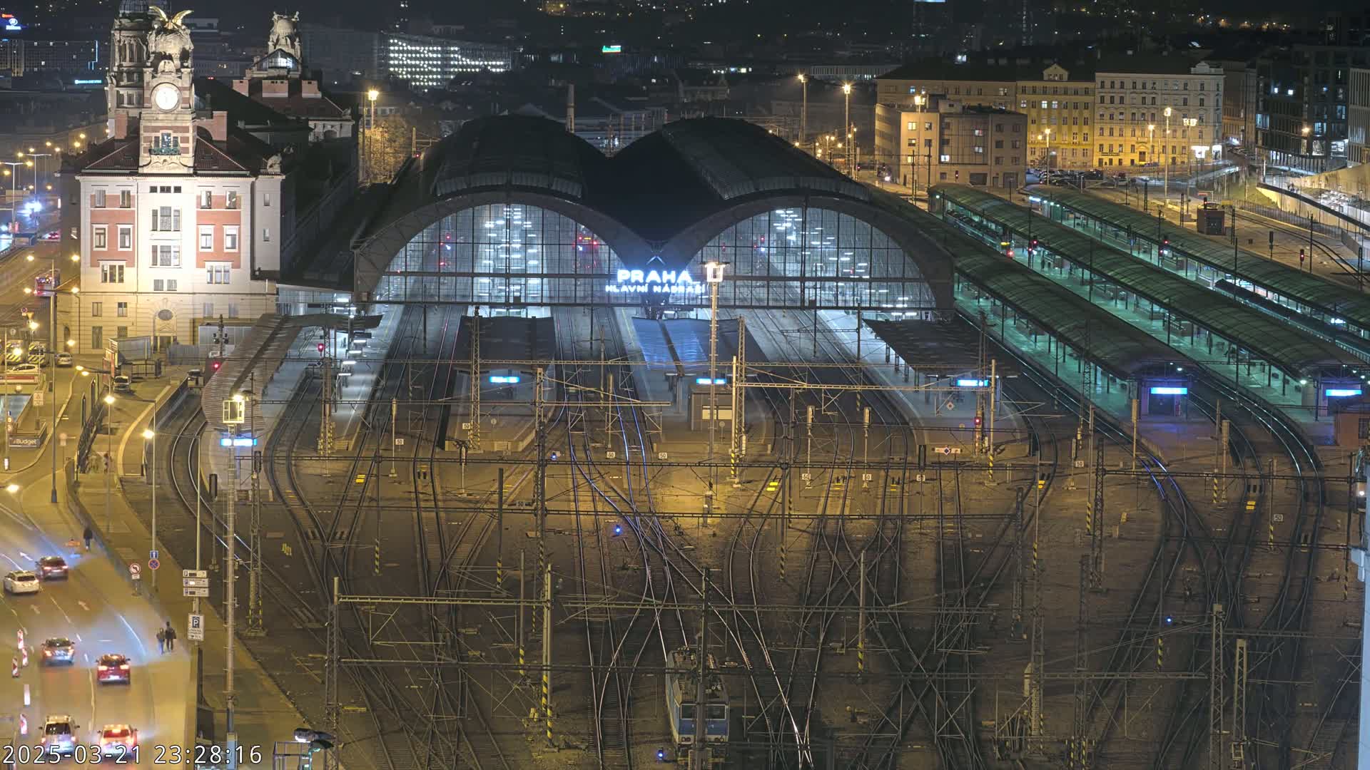 A nighttime aerial view of a large, illuminated train station with many tracks and platforms, surrounded by city streets and buildings.