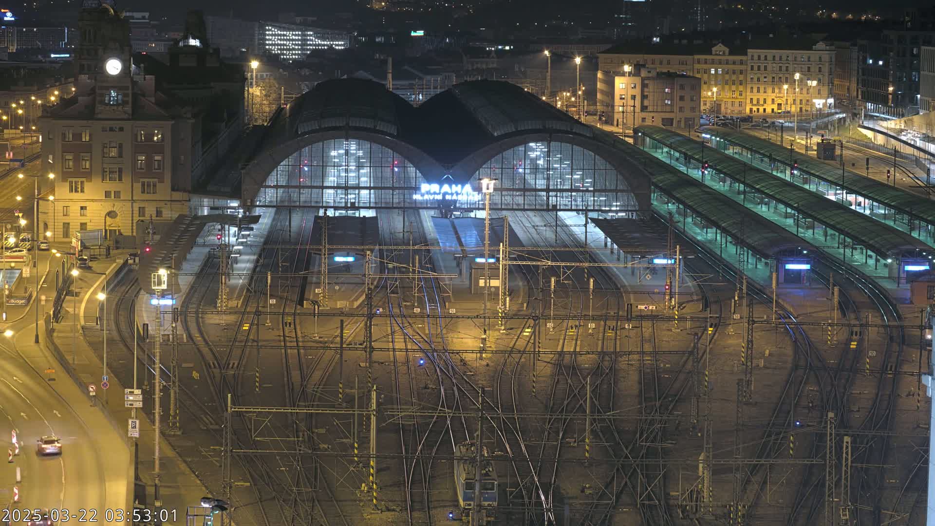 A large, illuminated train station at night, with numerous tracks and platforms extending from its main building, surrounded by city buildings under a dark sky.