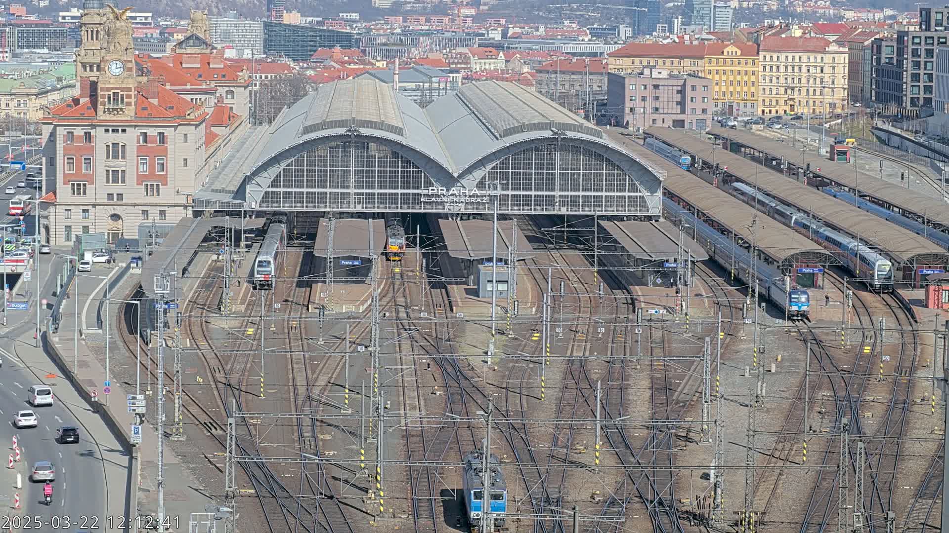 A large train station with many tracks and trains, surrounded by a city on a sunny day.