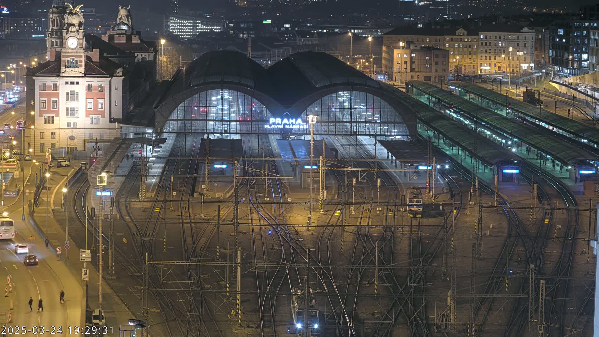 A nighttime, aerial view of a large, illuminated train station with numerous tracks and platforms, surrounded by city buildings.