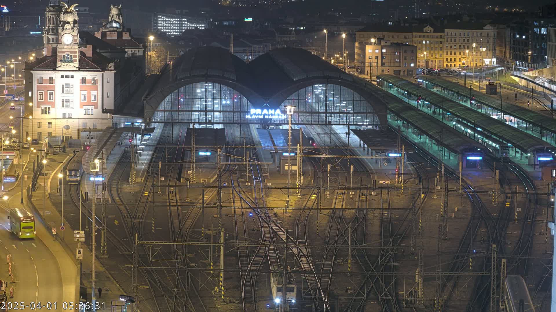 A large, illuminated train station at night with numerous tracks and platforms, surrounded by city buildings.