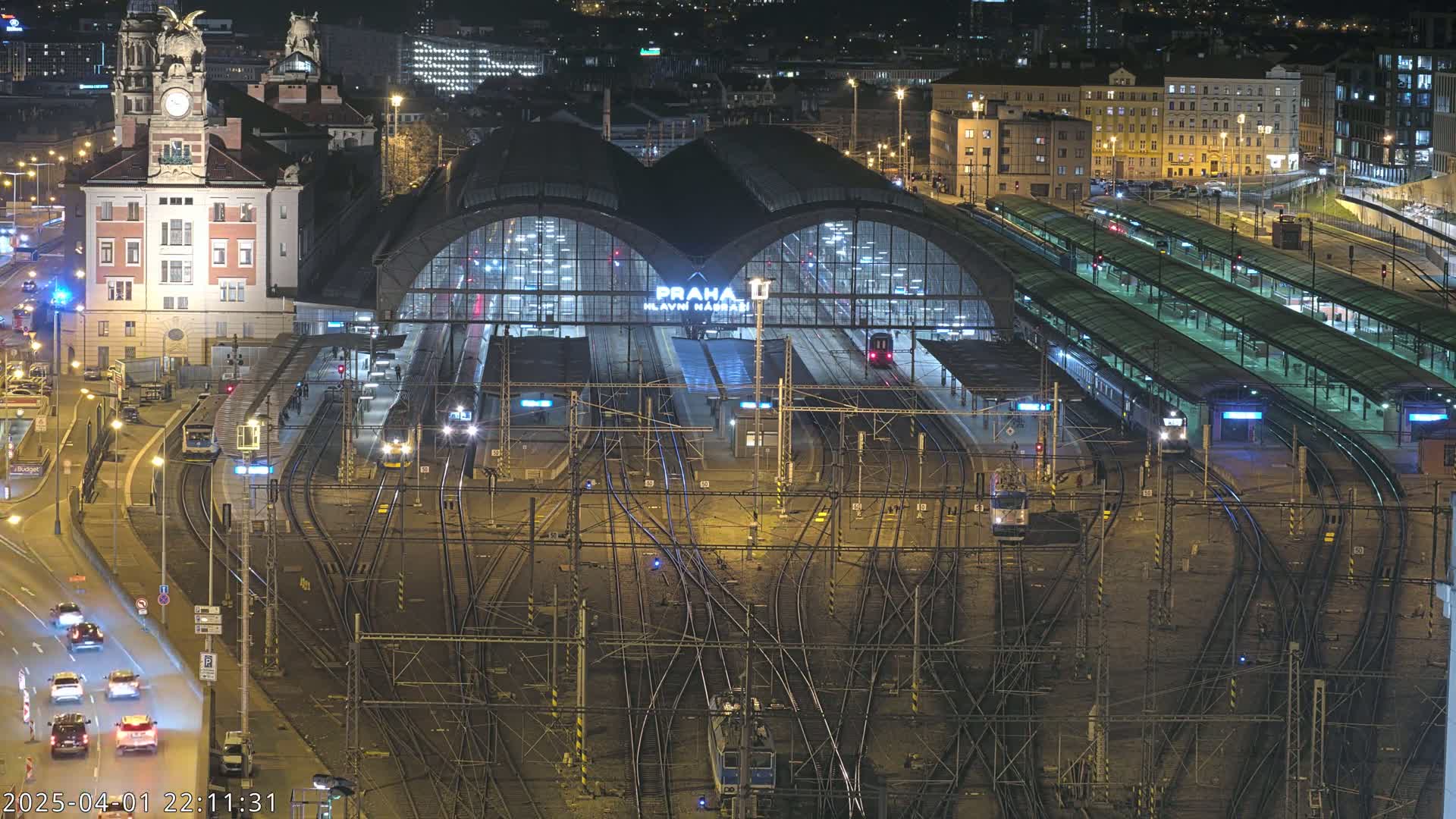 A nighttime aerial view of a large, busy train station with many tracks and platforms, illuminated by artificial lights, with cars visible on a road adjacent to the station.
