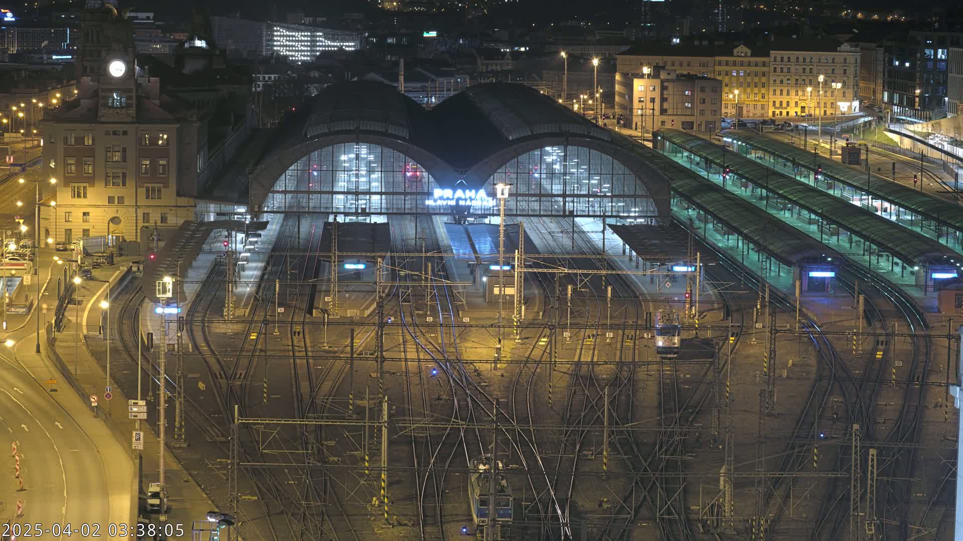 A large, illuminated train station at night, with numerous tracks and platforms extending from it, surrounded by city buildings.
