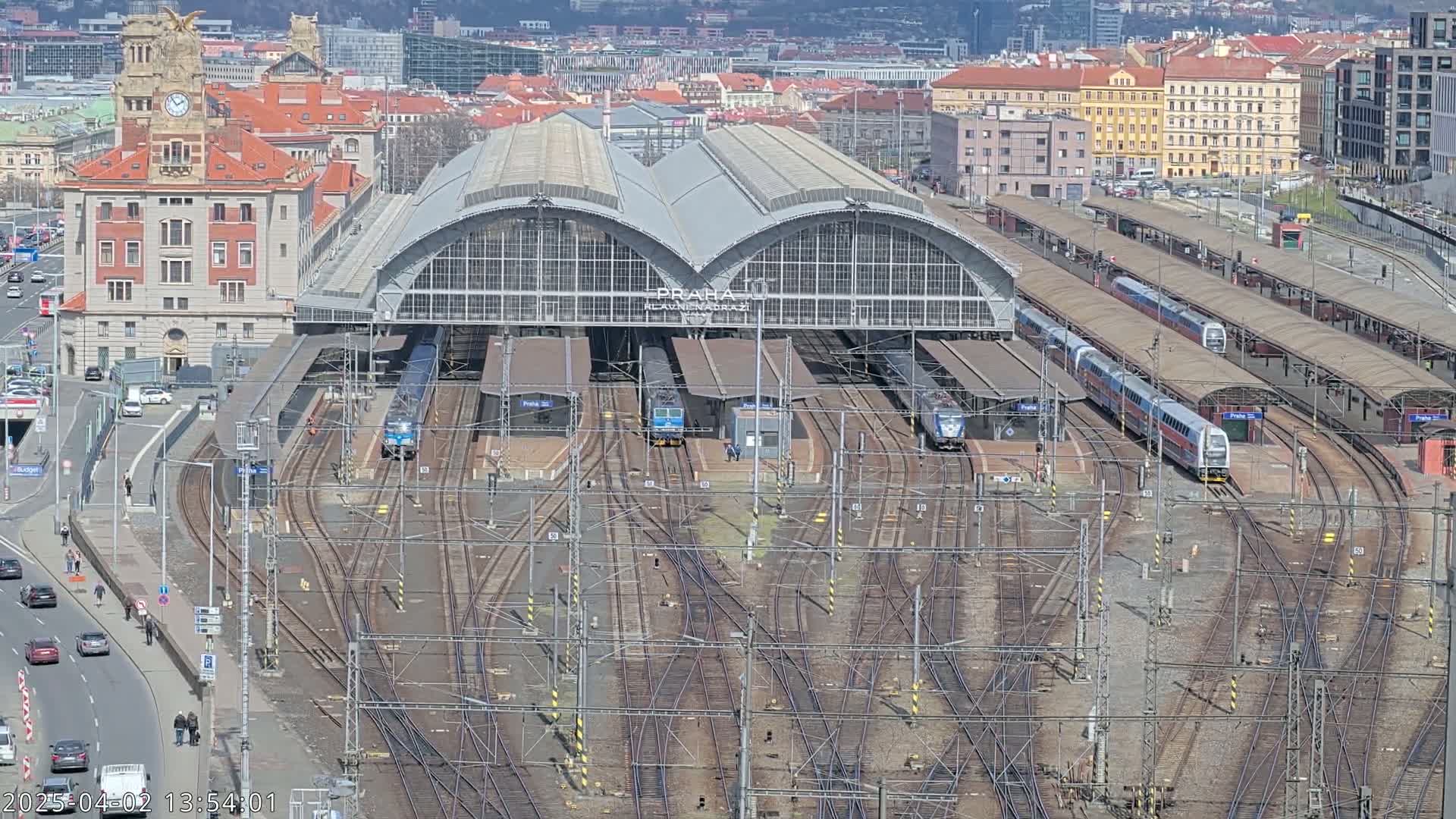 A large train station with numerous tracks and trains, under a sunny sky, is surrounded by a city.