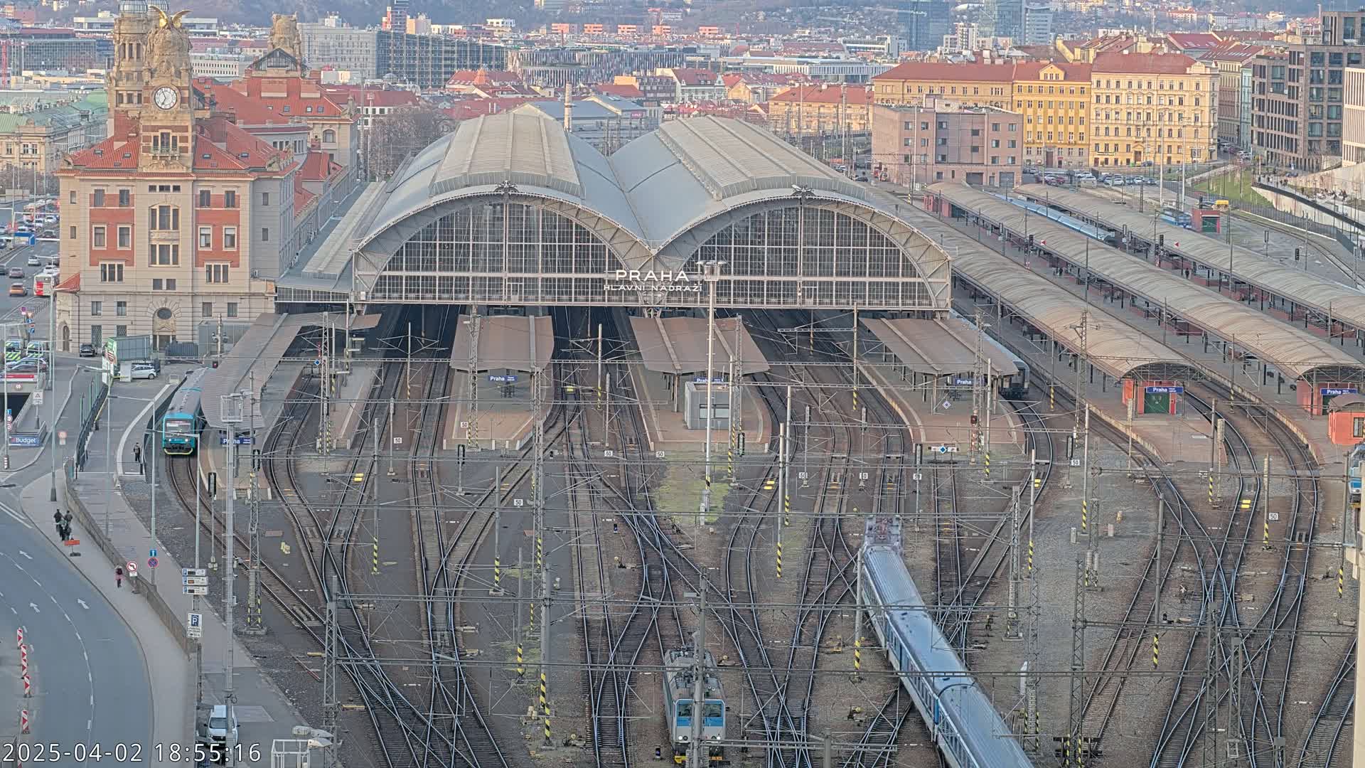A large train station with numerous tracks and platforms, surrounded by city buildings under a clear, daytime sky.