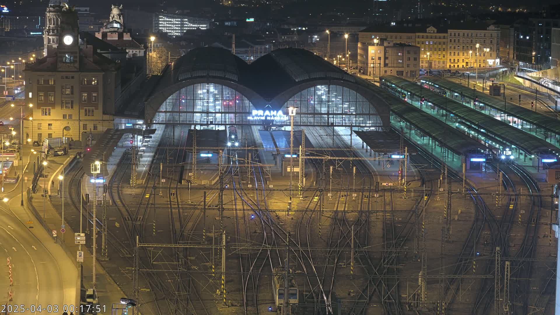 A nighttime aerial view of a large, illuminated train station with numerous tracks and platforms, surrounded by city buildings.