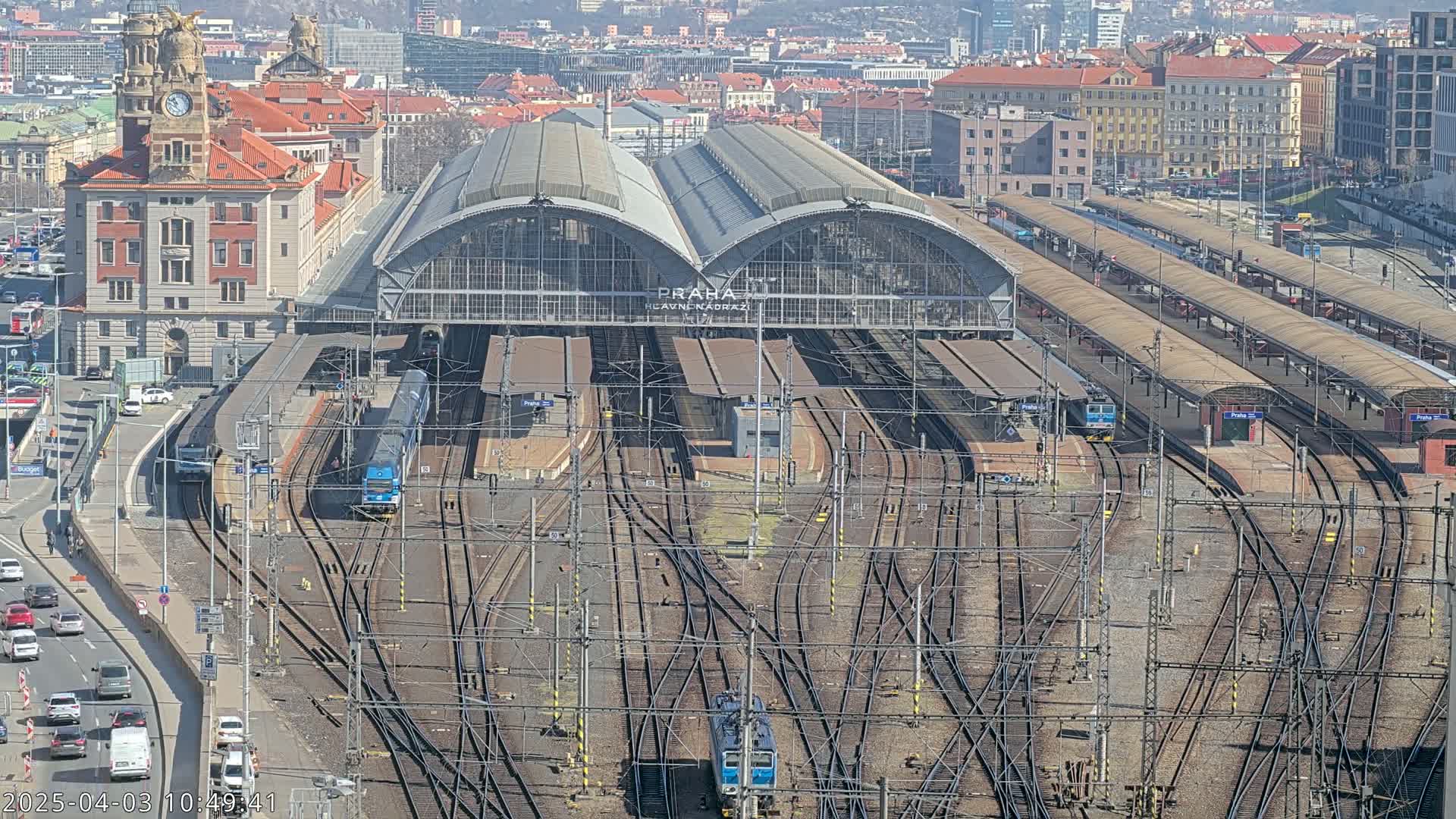 A large train station with numerous tracks and trains, under a sunny sky, is viewed from above.