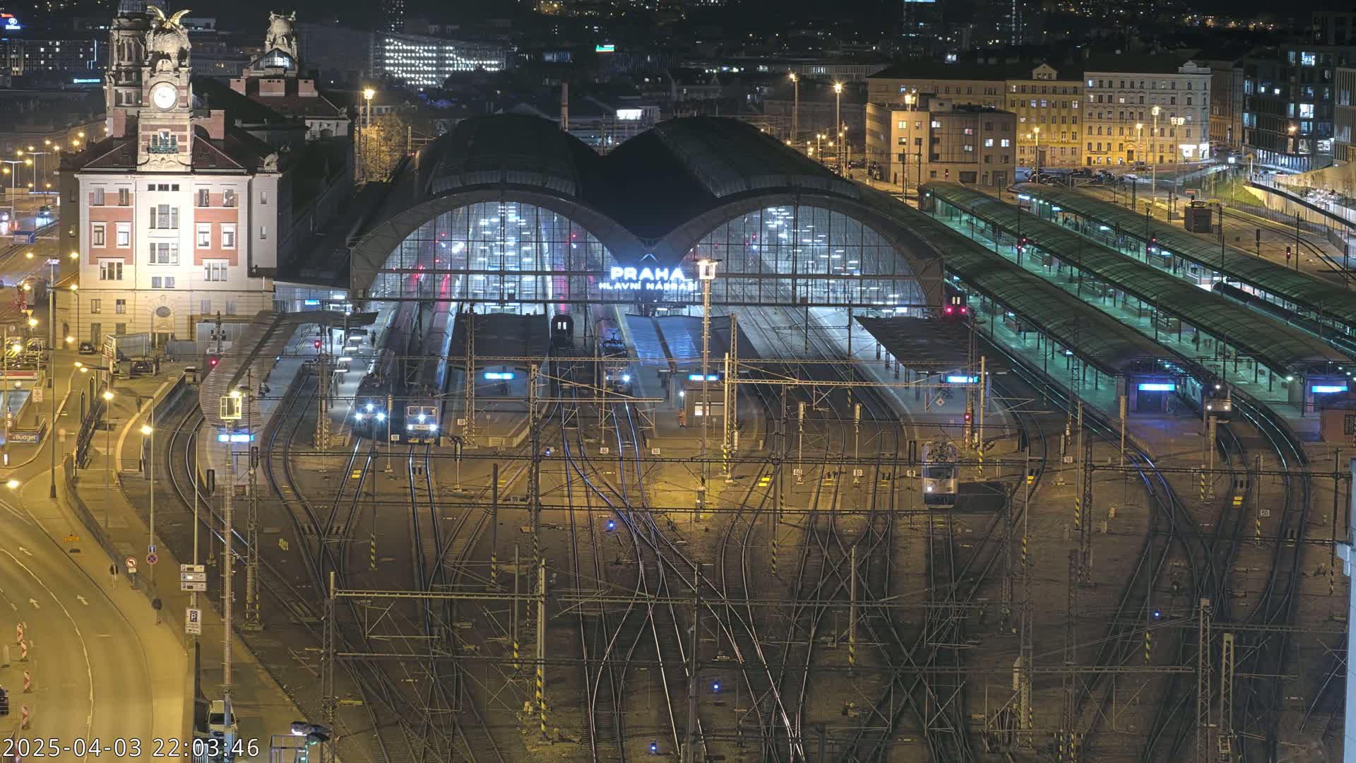 A nighttime, aerial view of a large, illuminated train station with many tracks and platforms, surrounded by city buildings.