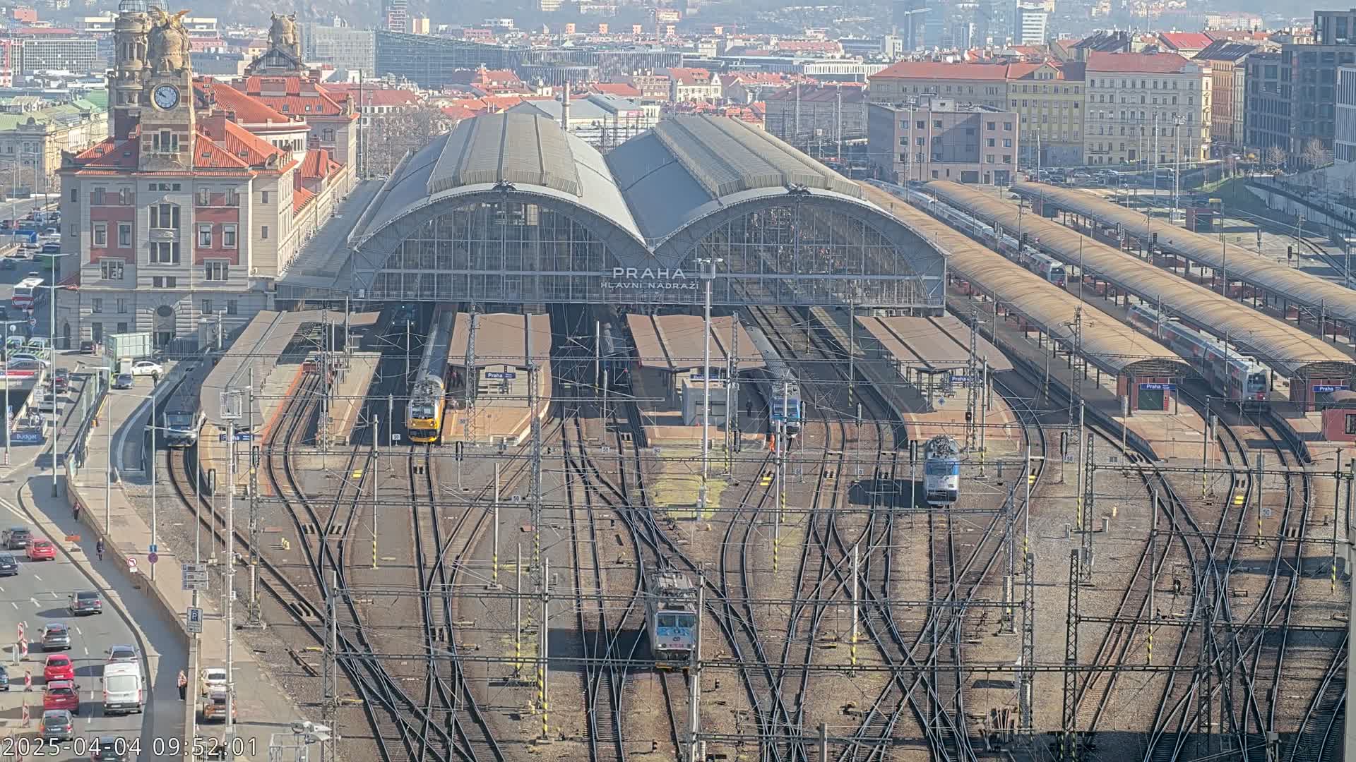 A large train station with many tracks and trains, under a sunny sky, is surrounded by city buildings.