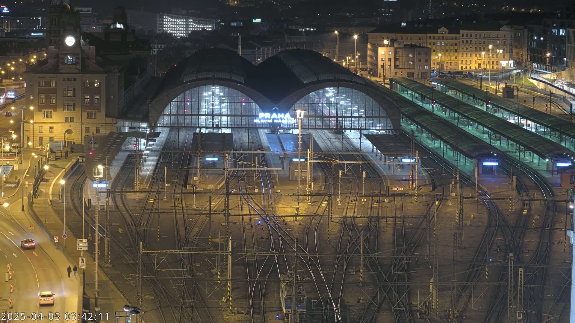 A large, illuminated train station at night, with numerous tracks and platforms extending from it, surrounded by city buildings and streets.