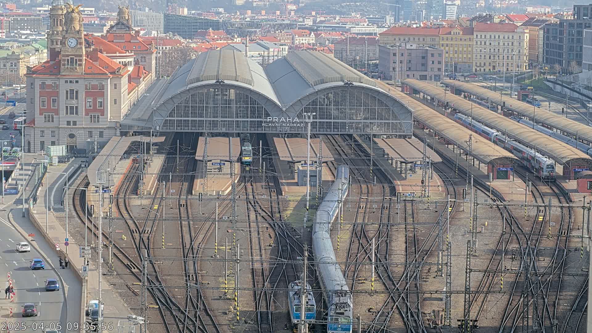 A large train station with many tracks and trains, surrounded by city buildings under a clear sky.