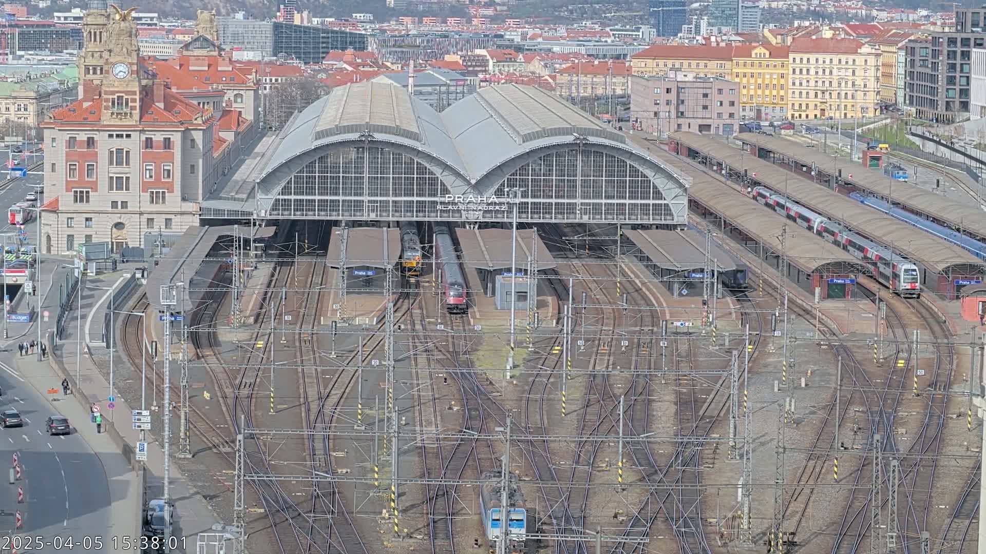 A large train station with many tracks and several trains, under a sunny sky, is surrounded by a city.