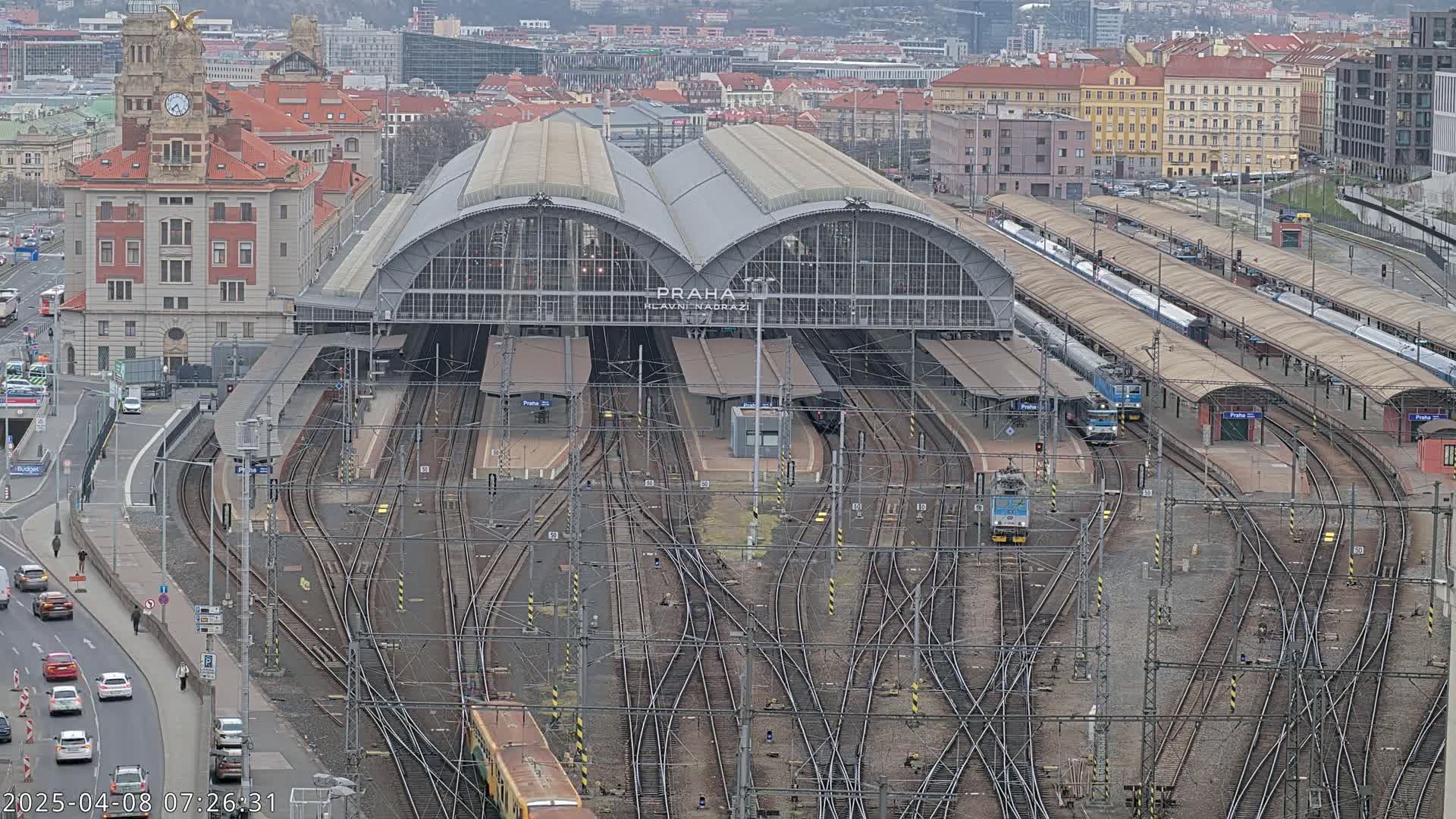 A large train station with numerous tracks and several trains, surrounded by city buildings under an overcast sky.
