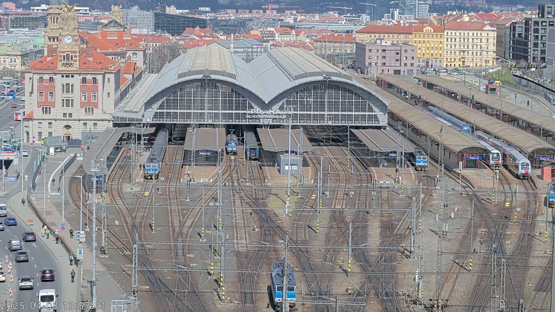 A large train station with numerous tracks and several trains, situated in a city under a sunny sky.