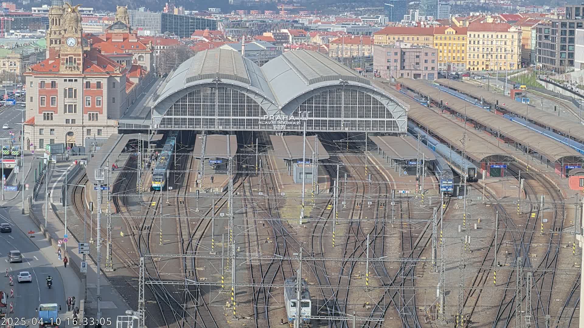 A large train station with numerous tracks and trains, under a sunny sky, is surrounded by a city.