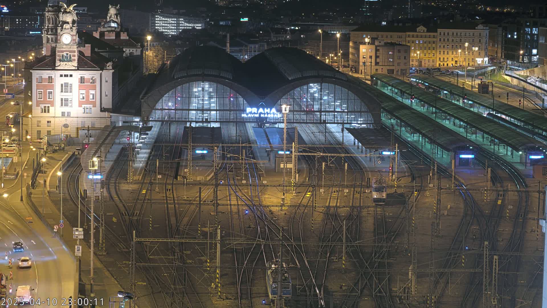 A brightly lit train station at night, featuring numerous train tracks and platforms, with a large building in the background.
