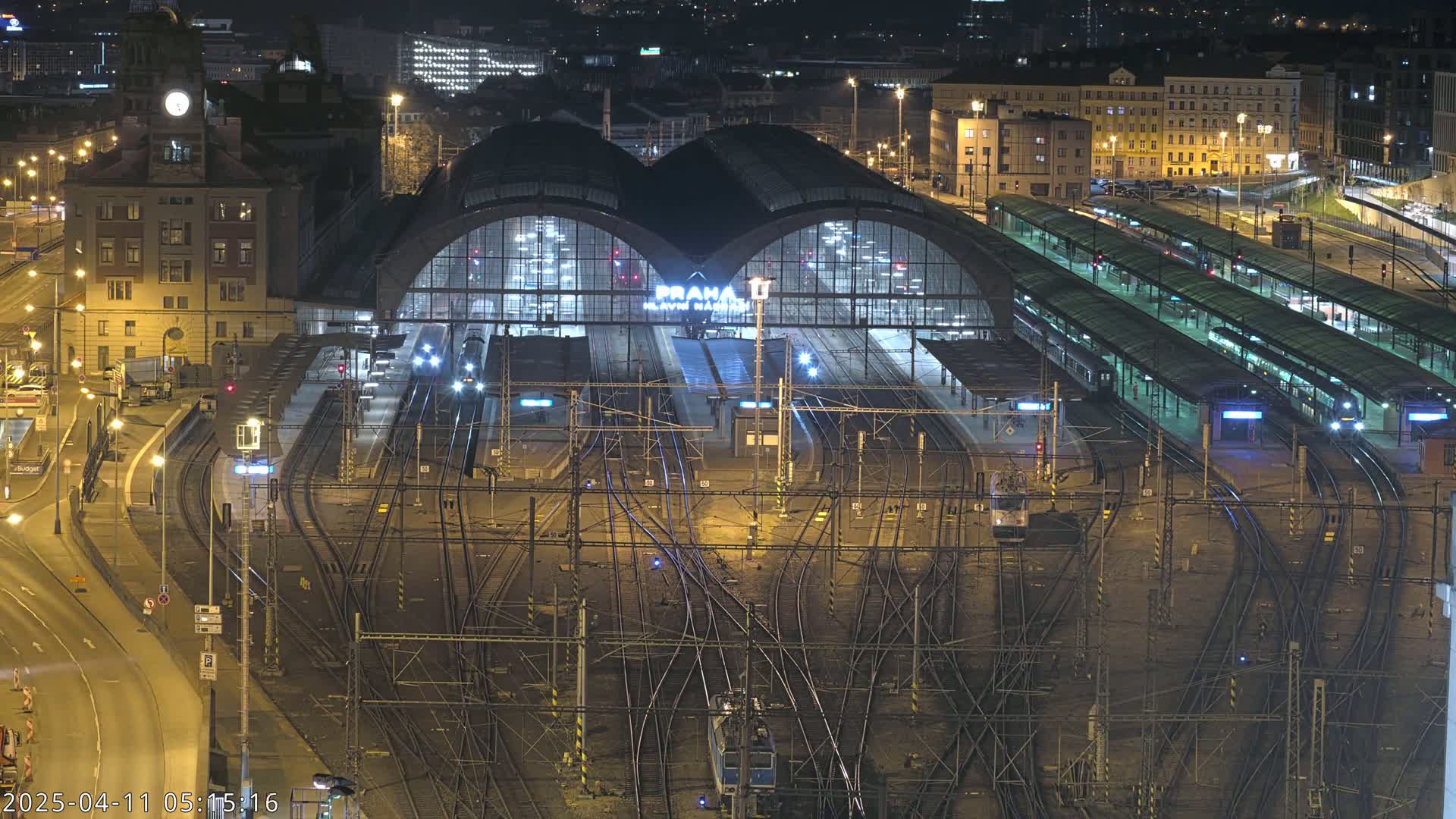 A large, illuminated train station at night with numerous train tracks and platforms, surrounded by city buildings.