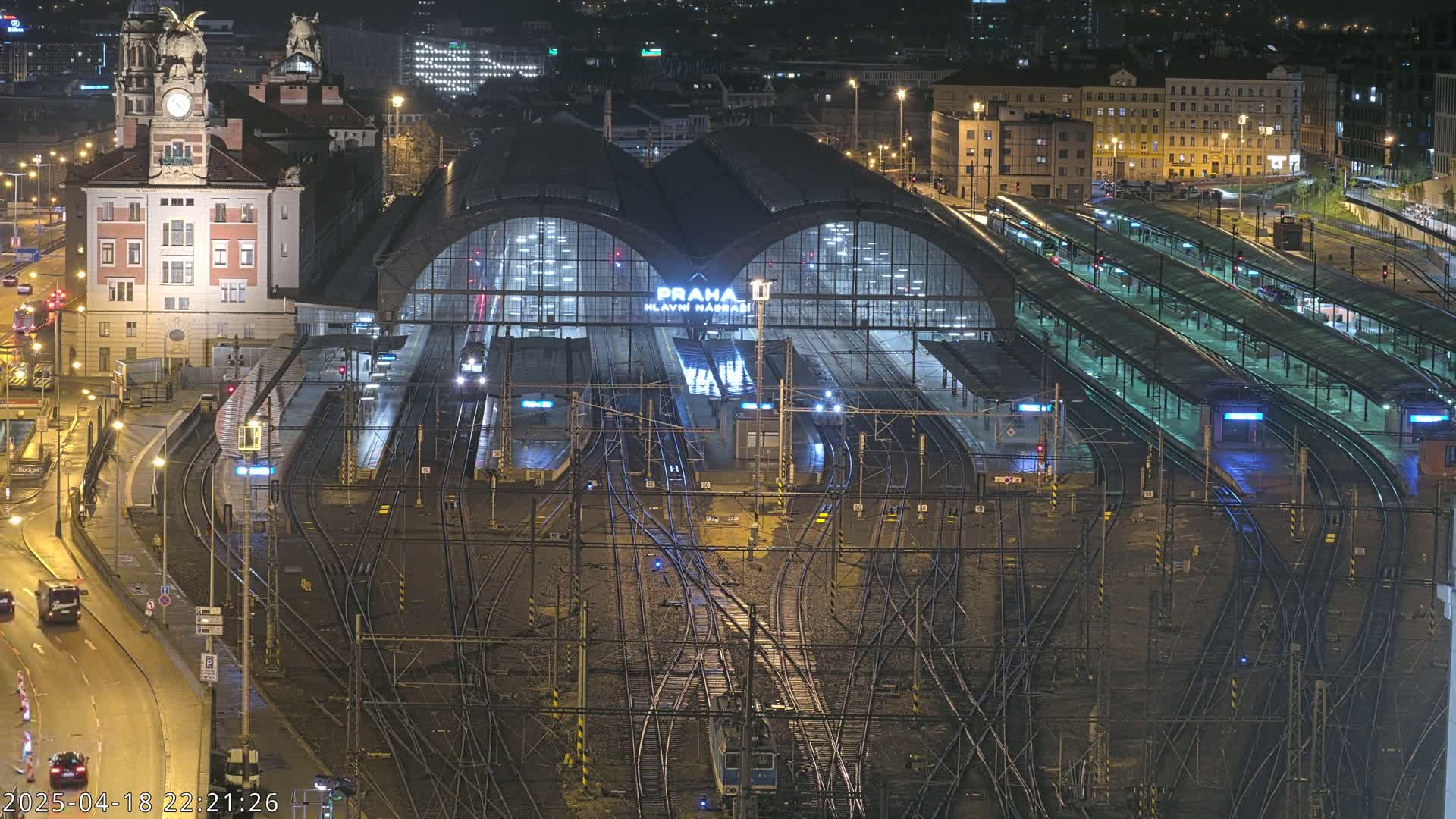 A nighttime aerial view of a large, illuminated train station with numerous tracks and platforms, surrounded by city buildings.