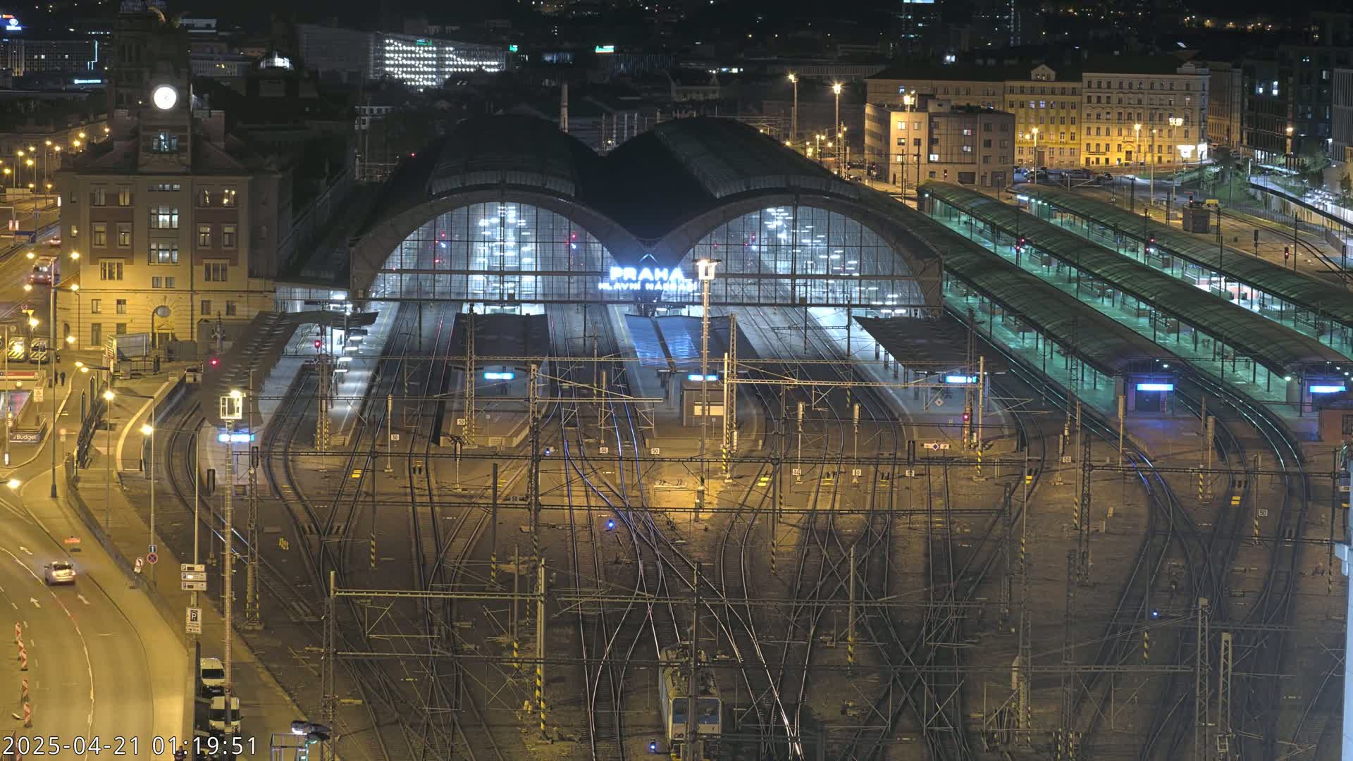 A large, illuminated train station at night, surrounded by a complex network of tracks and adjacent buildings.