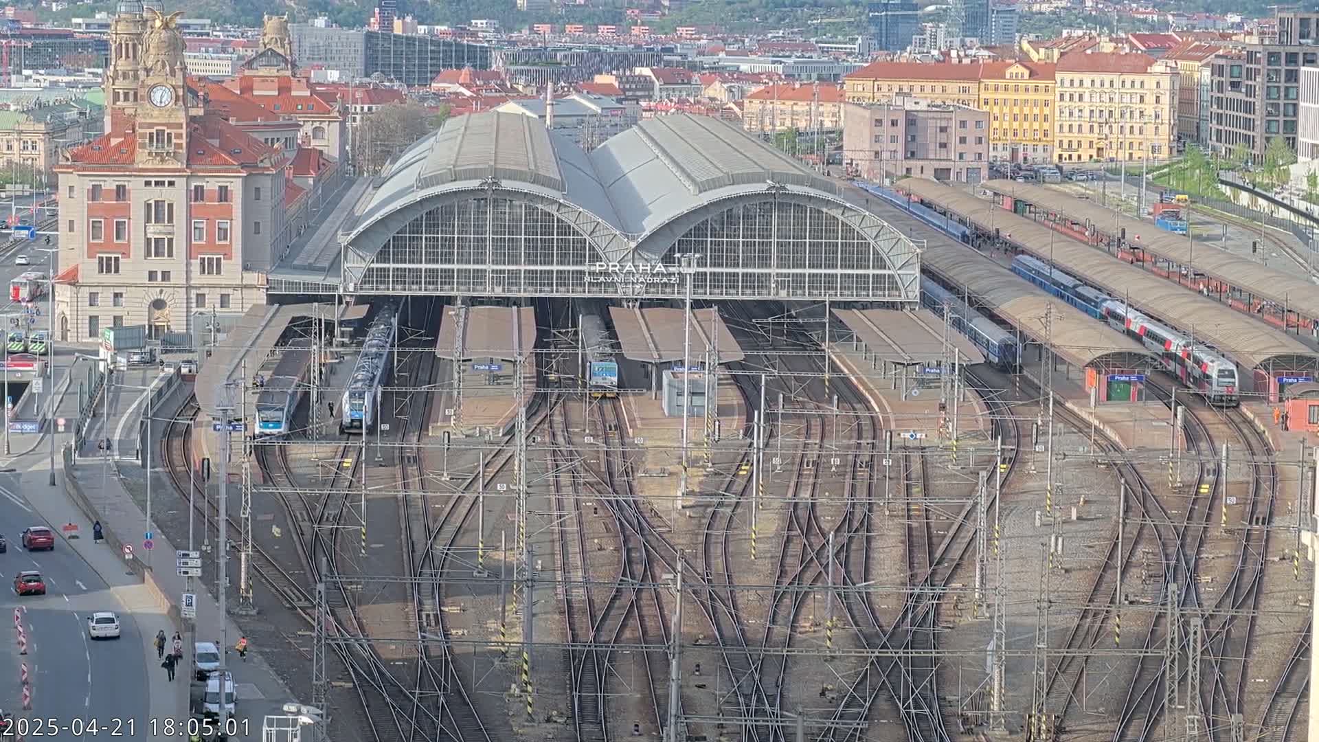 A large train station with multiple tracks and trains, surrounded by city buildings under a partly sunny sky.