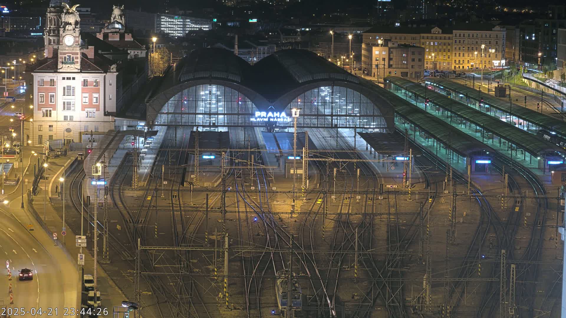 A brightly lit train station at night, with numerous train tracks and platforms extending from a large central building, surrounded by city buildings.
