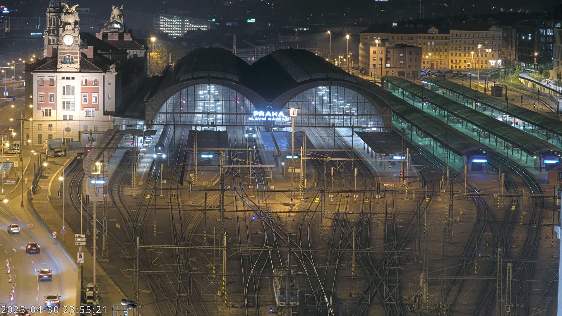 A nighttime, high-angle view of a large train station with numerous tracks and platforms, and a building with a clock tower adjacent to it.