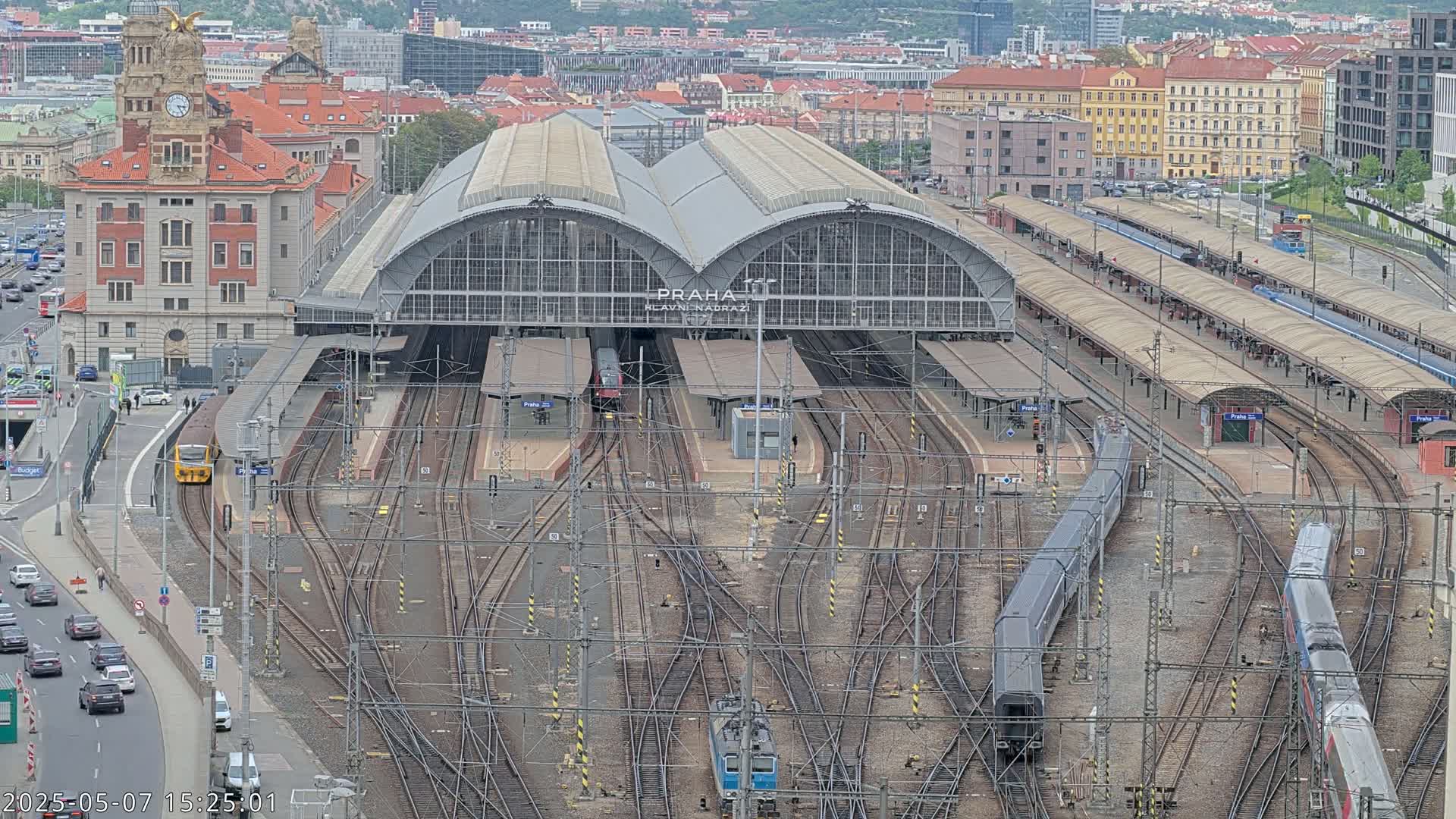 A large train station with numerous tracks and trains, surrounded by city buildings under a cloudy sky.