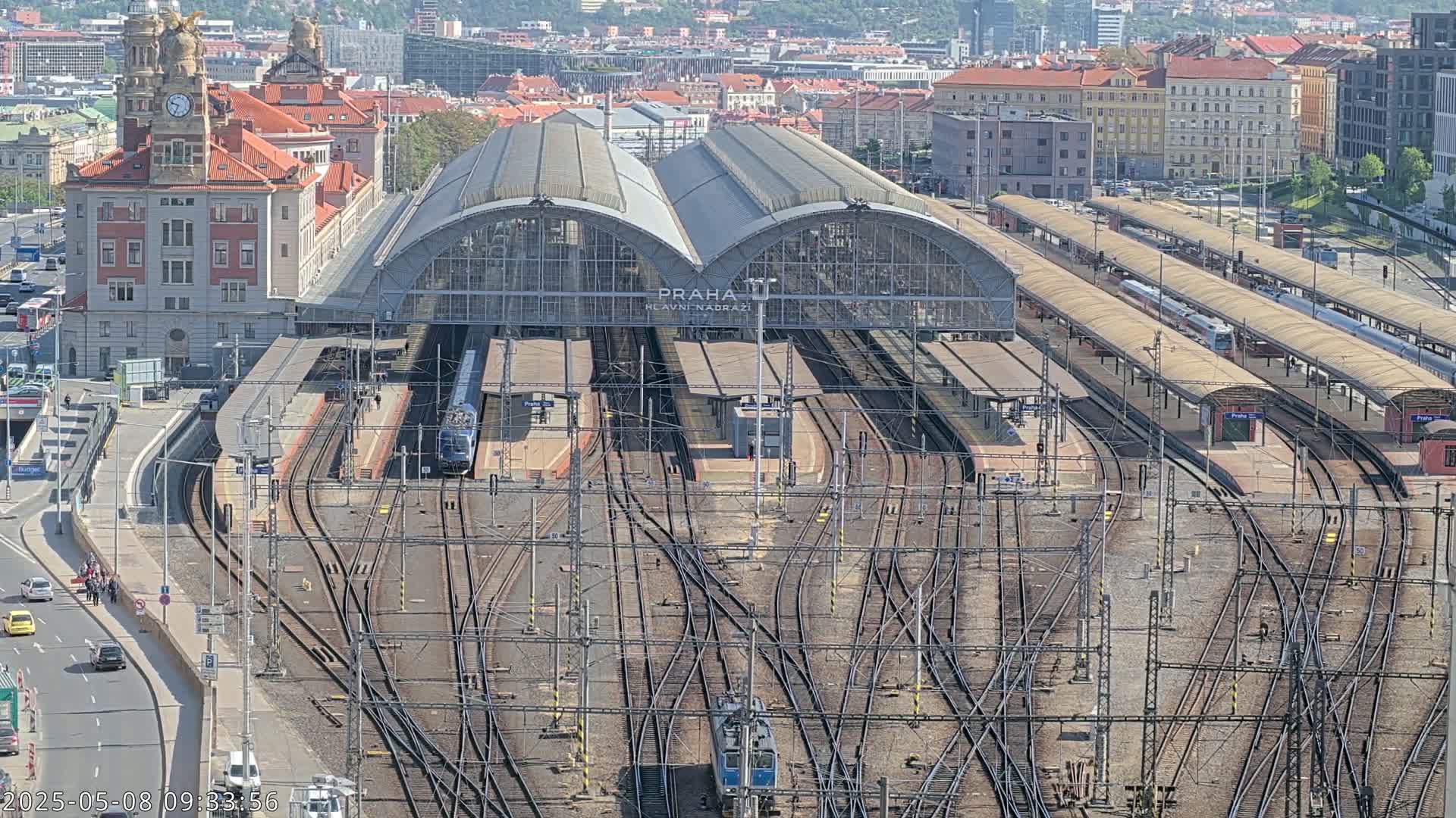 A large train station with numerous tracks and platforms, surrounded by city buildings on a sunny day.