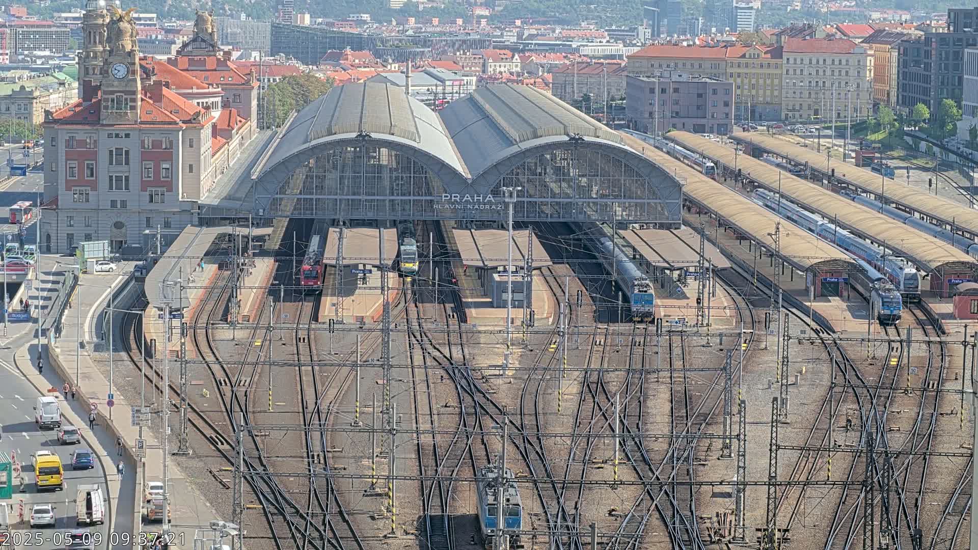 A large train station with numerous tracks and trains, under a sunny sky, is surrounded by city buildings.