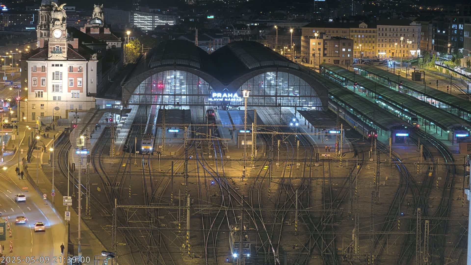 A nighttime aerial view of a large, illuminated train station with numerous tracks and trains, surrounded by city buildings and streets.