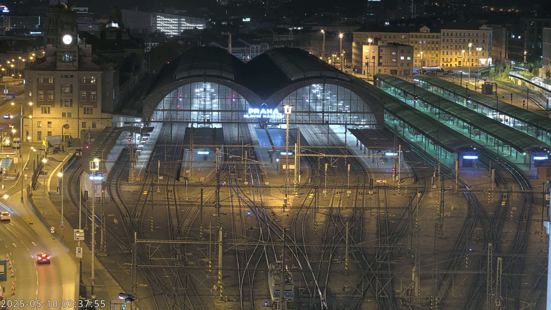 A nighttime aerial view of a large, illuminated train station with numerous tracks and platforms, surrounded by city buildings.