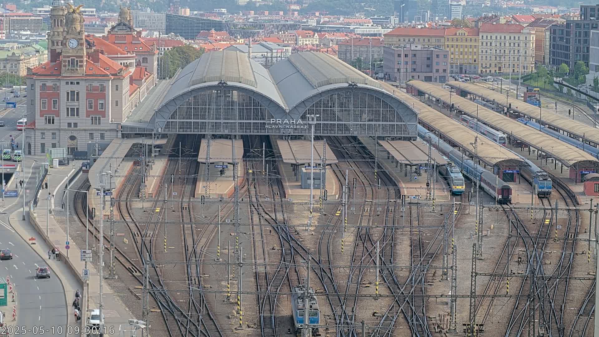 A large train station with numerous tracks and trains, a large station building, and surrounding city buildings is seen on a clear day.