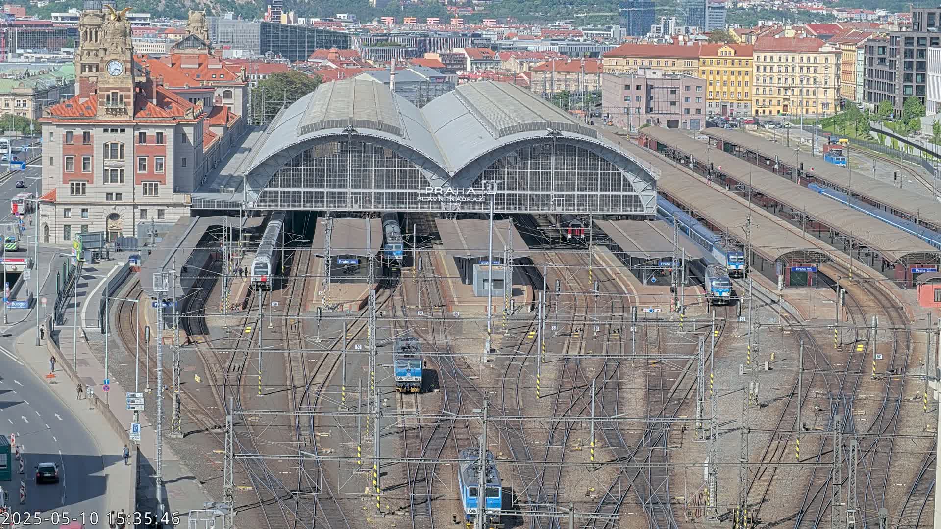 A large train station with numerous tracks and trains, under a sunny sky, is surrounded by city buildings.