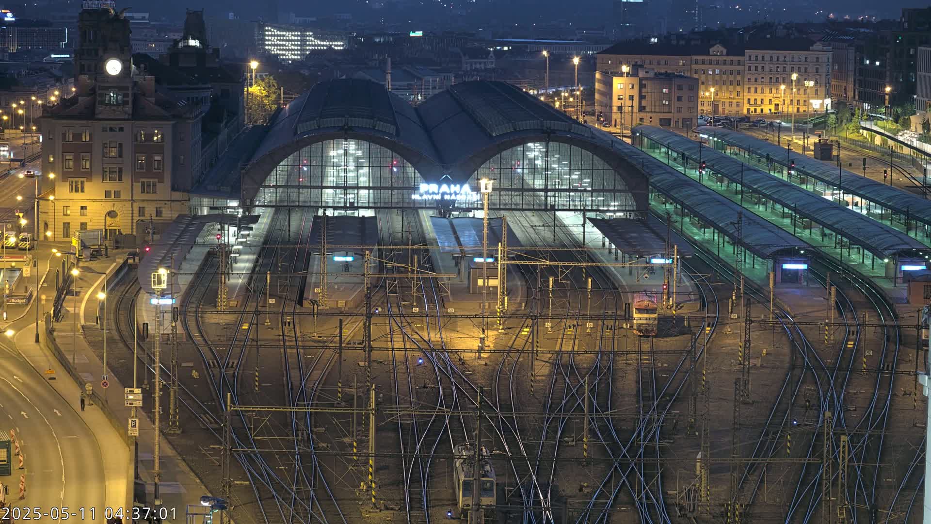A large, illuminated train station at night, surrounded by numerous train tracks and platforms, with city buildings visible in the background under a dark sky.
