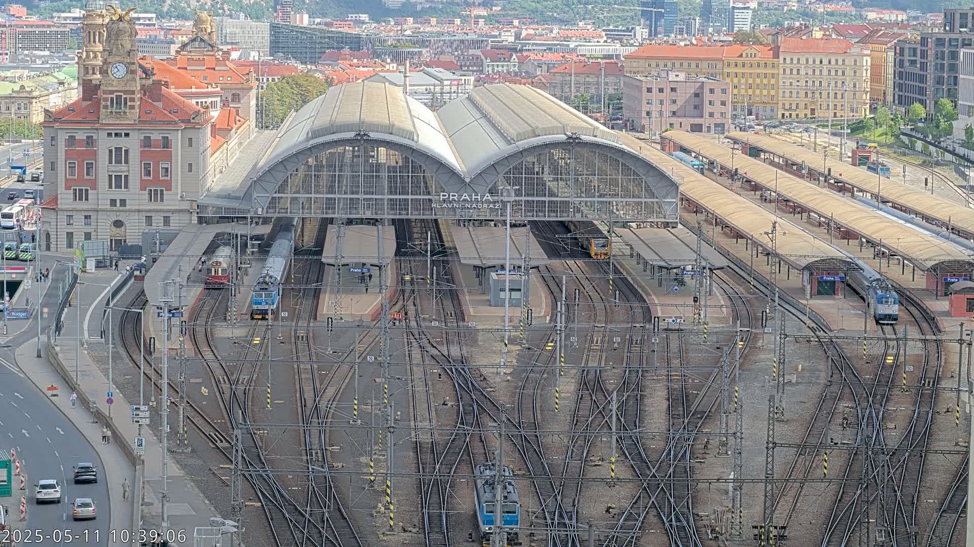 A large train station with numerous tracks and trains, surrounded by city buildings on a sunny day.