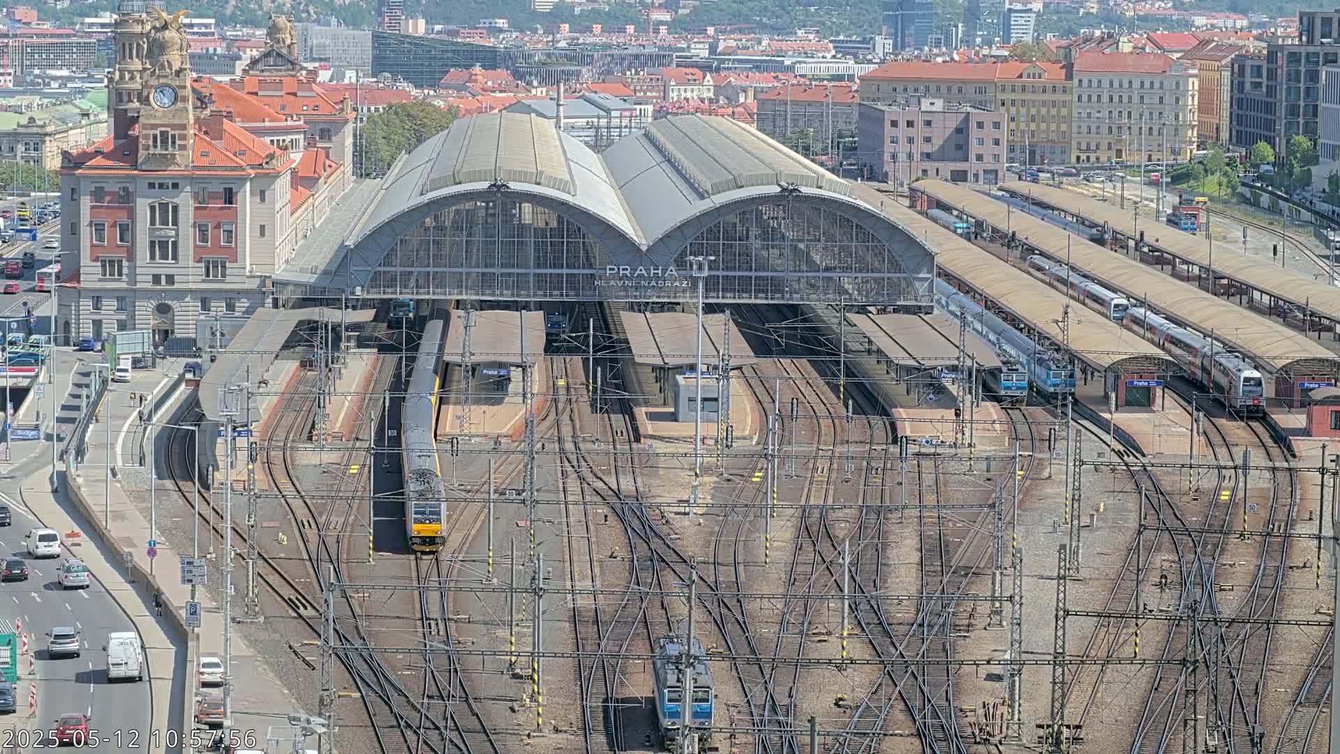 A large train station with numerous tracks and trains, under a sunny sky, is viewed from above.