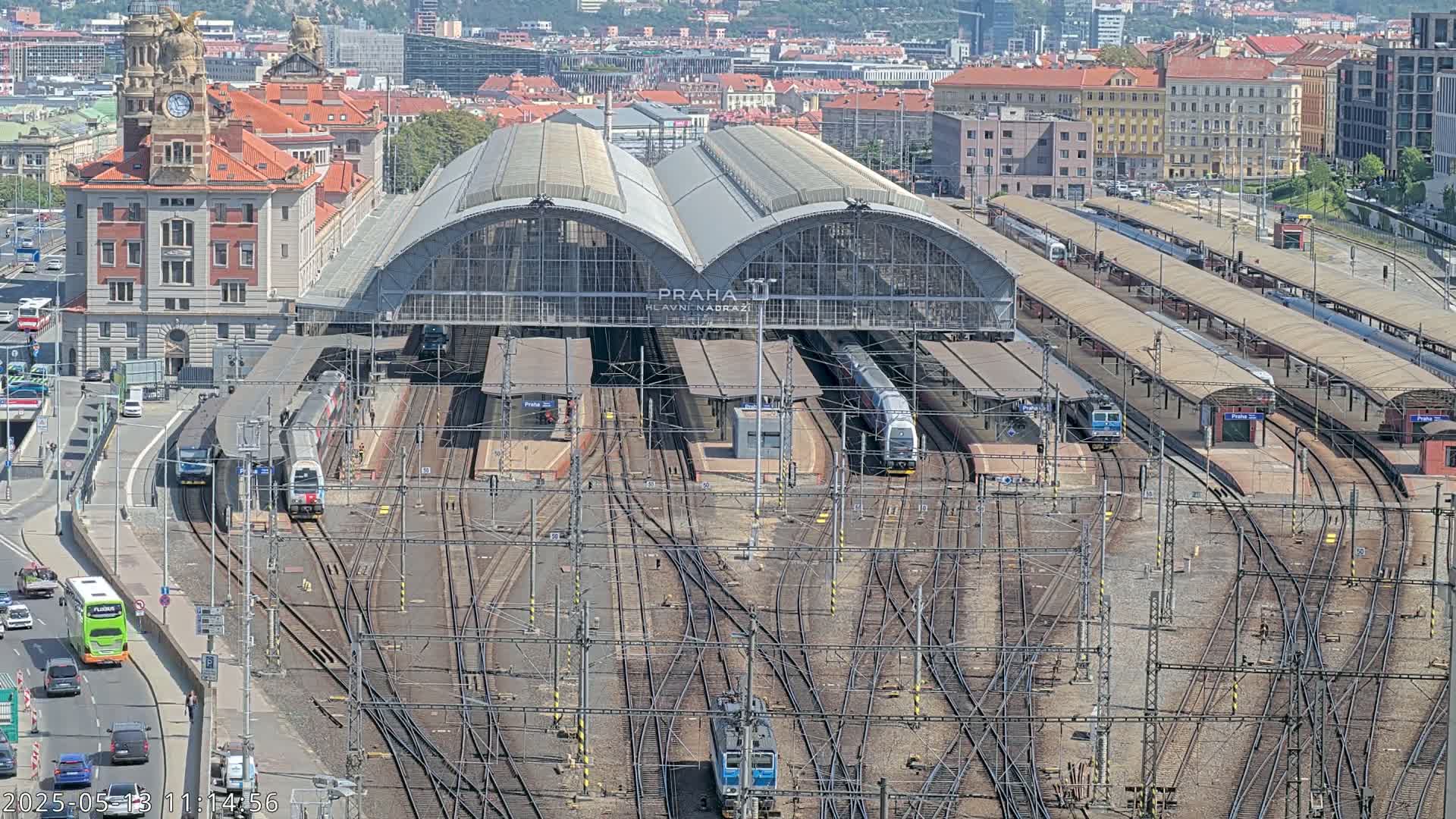 A large train station with numerous tracks and trains, surrounded by city buildings on a sunny day.
