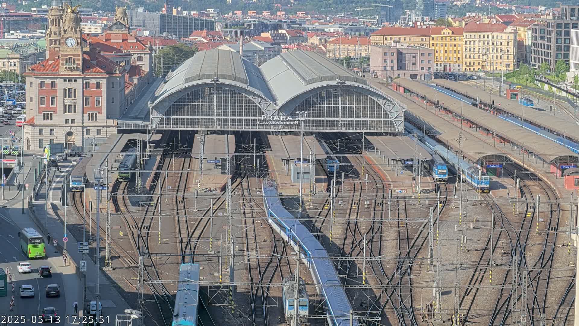 A large train station with numerous tracks and trains, under a sunny sky, is surrounded by city buildings.