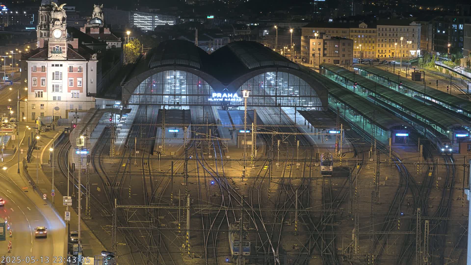 A brightly lit train station at night, with numerous tracks and platforms extending from a large central building, surrounded by city streets and buildings.