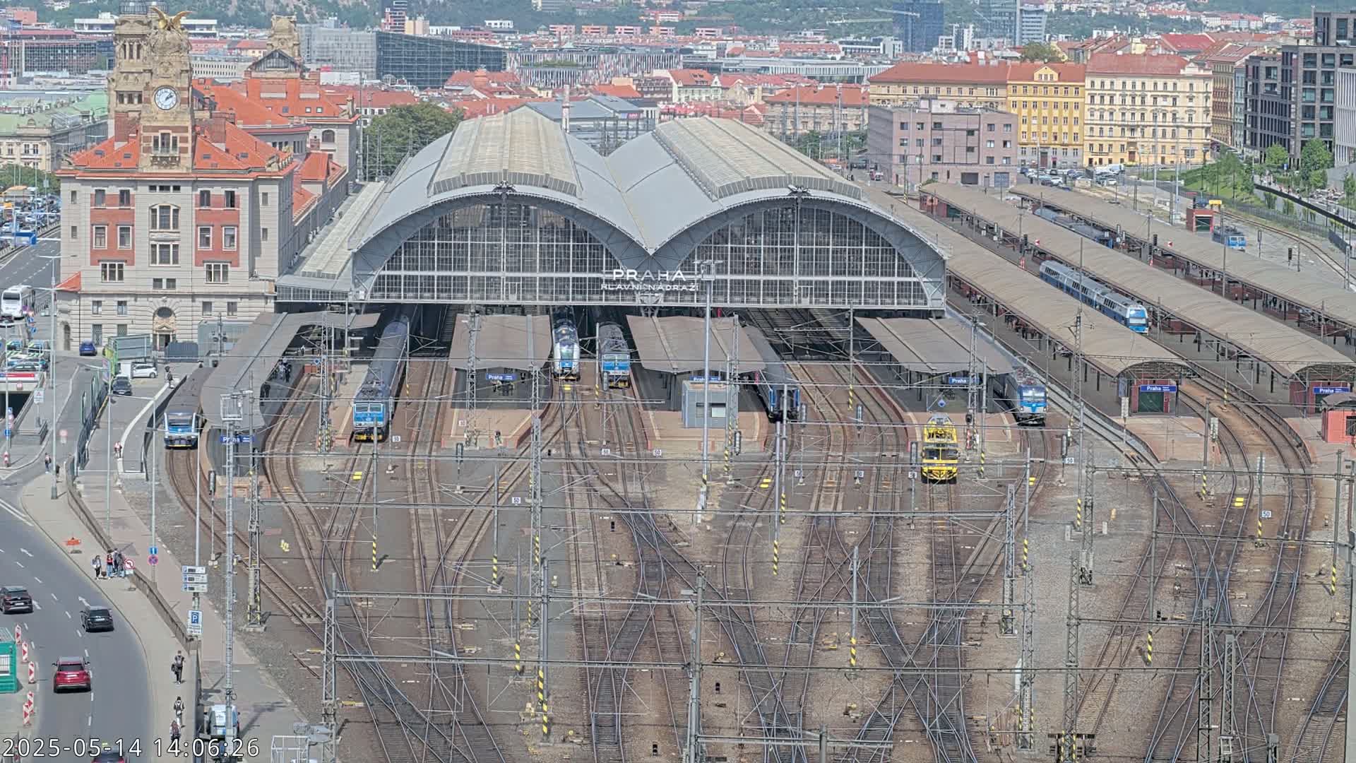 A large train station with many tracks and several trains, under a sunny sky, is surrounded by city buildings.