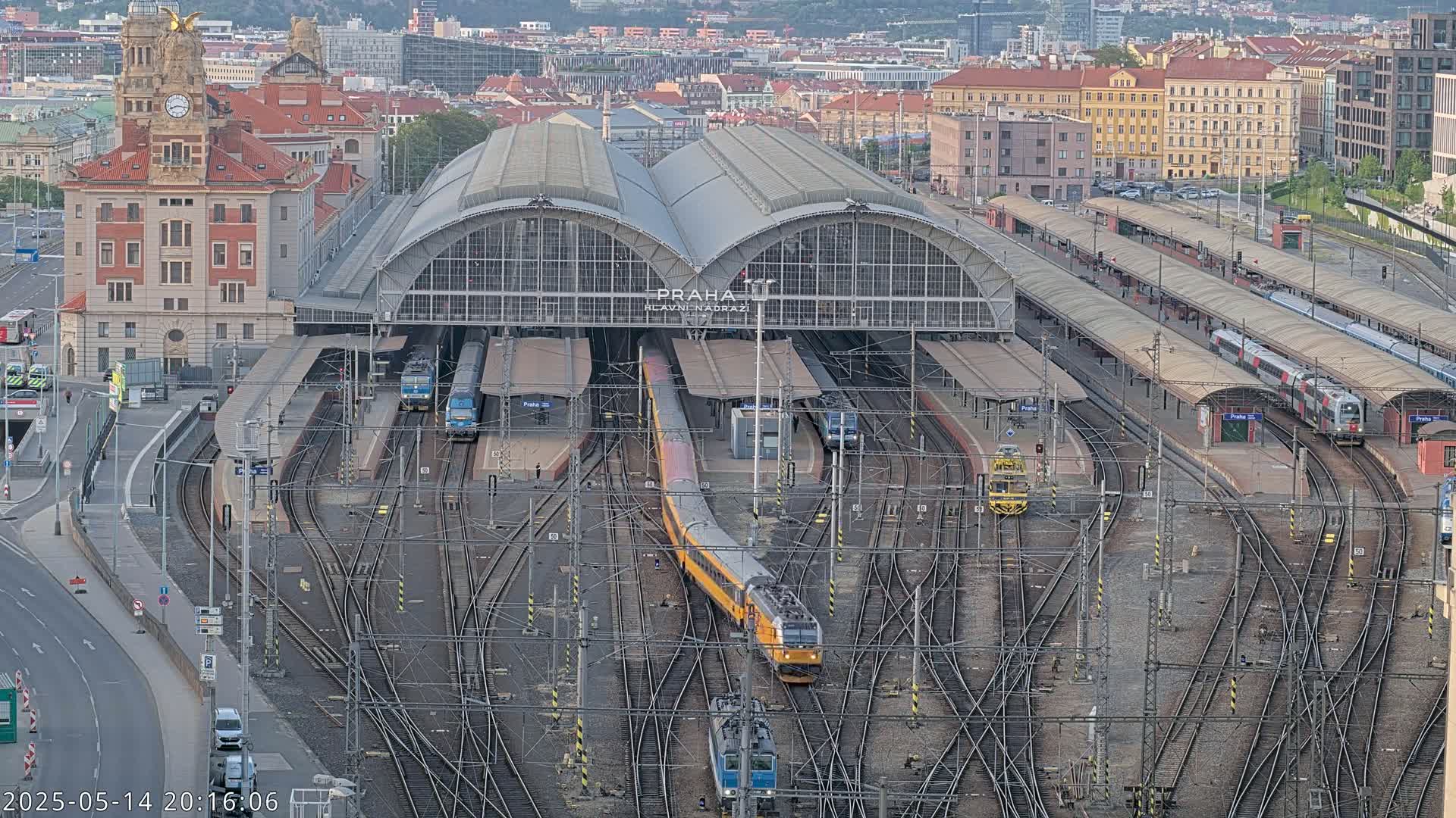 A large train station with numerous tracks and trains, some of which are yellow and others are silver, is situated in a city under a mostly clear sky.