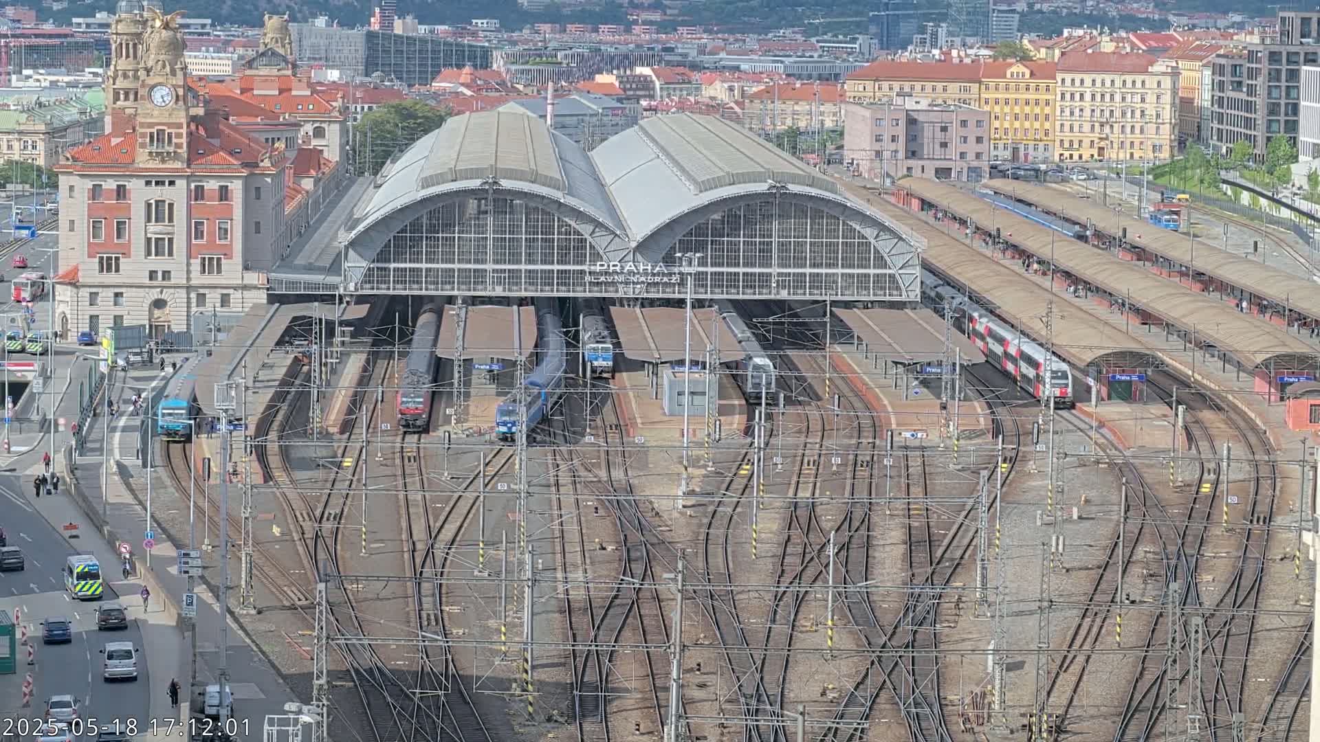 A large train station with multiple tracks and trains, surrounded by city buildings on a sunny day.