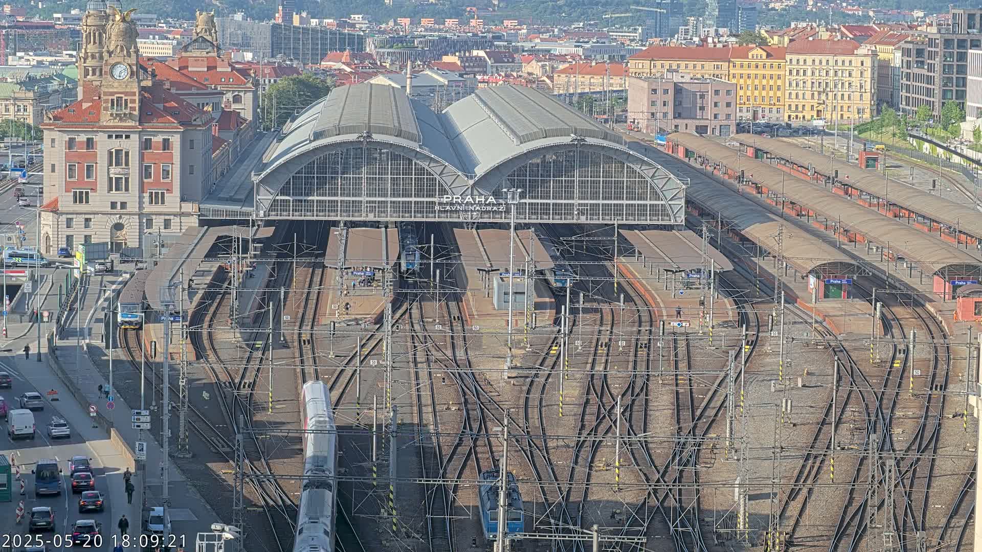 A large train station with numerous tracks and trains, surrounded by city buildings under a mostly sunny sky.