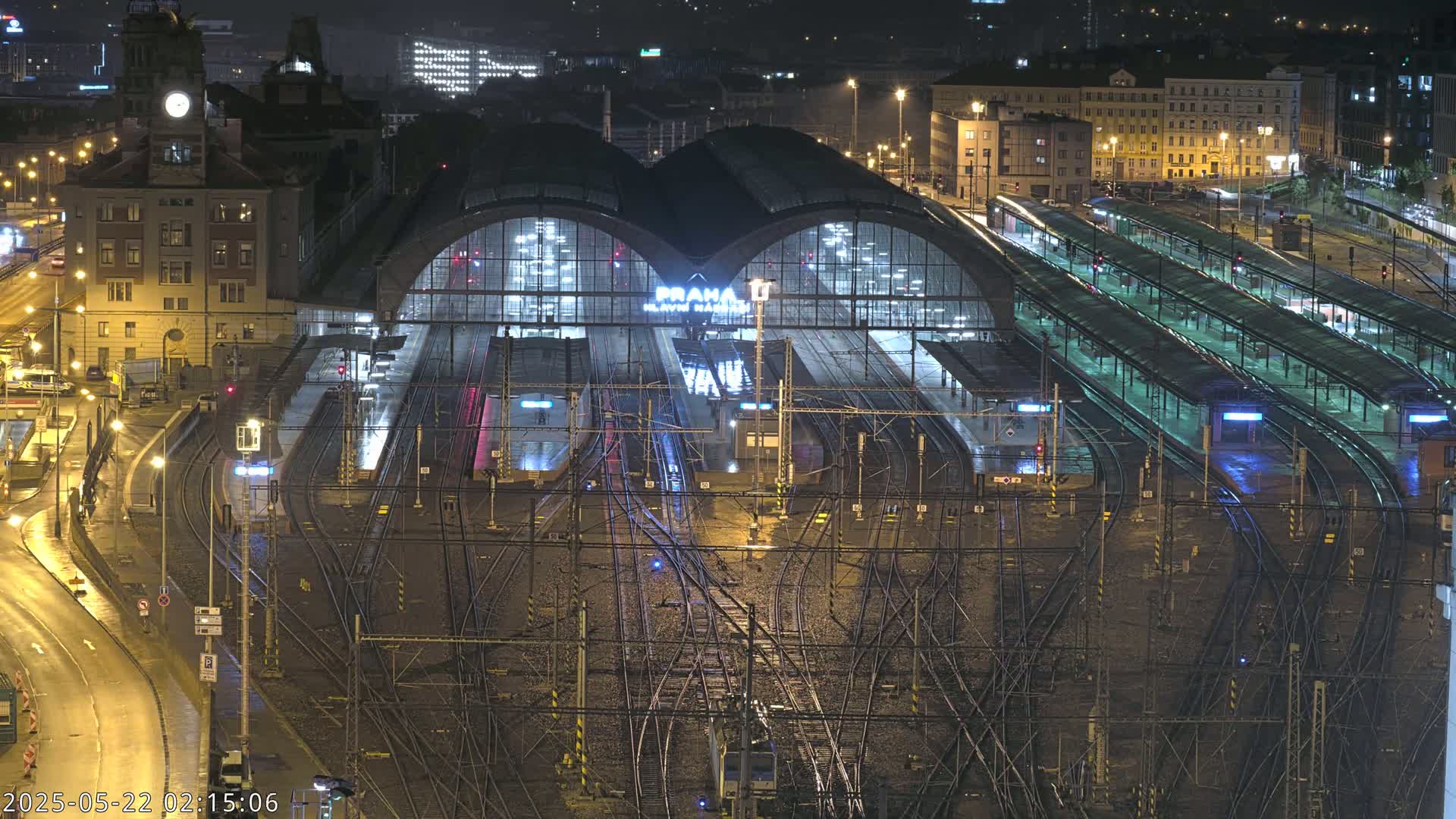 A large, illuminated train station at night, with numerous tracks and platforms under a wet, dark sky.