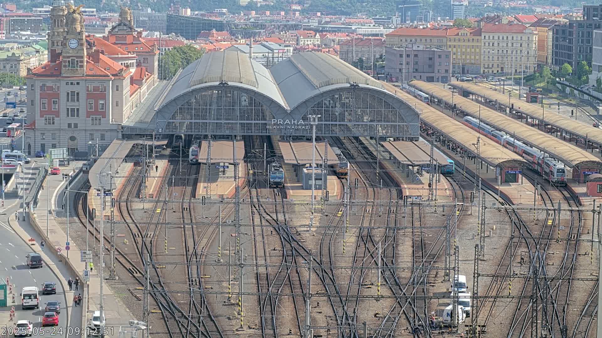 A large train station with numerous tracks and trains, surrounded by city buildings under a sunny sky.