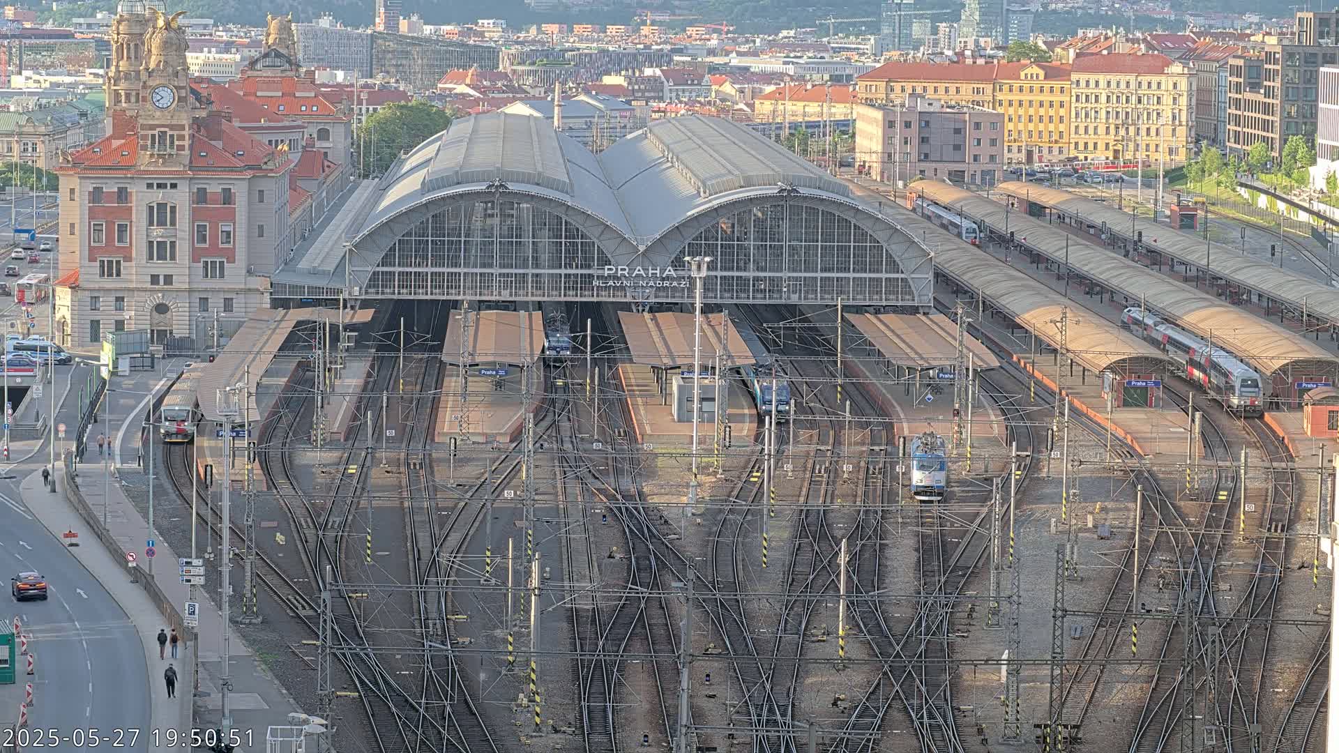 A large, arched train station with numerous tracks and trains sits in a city under a clear, daytime sky.
